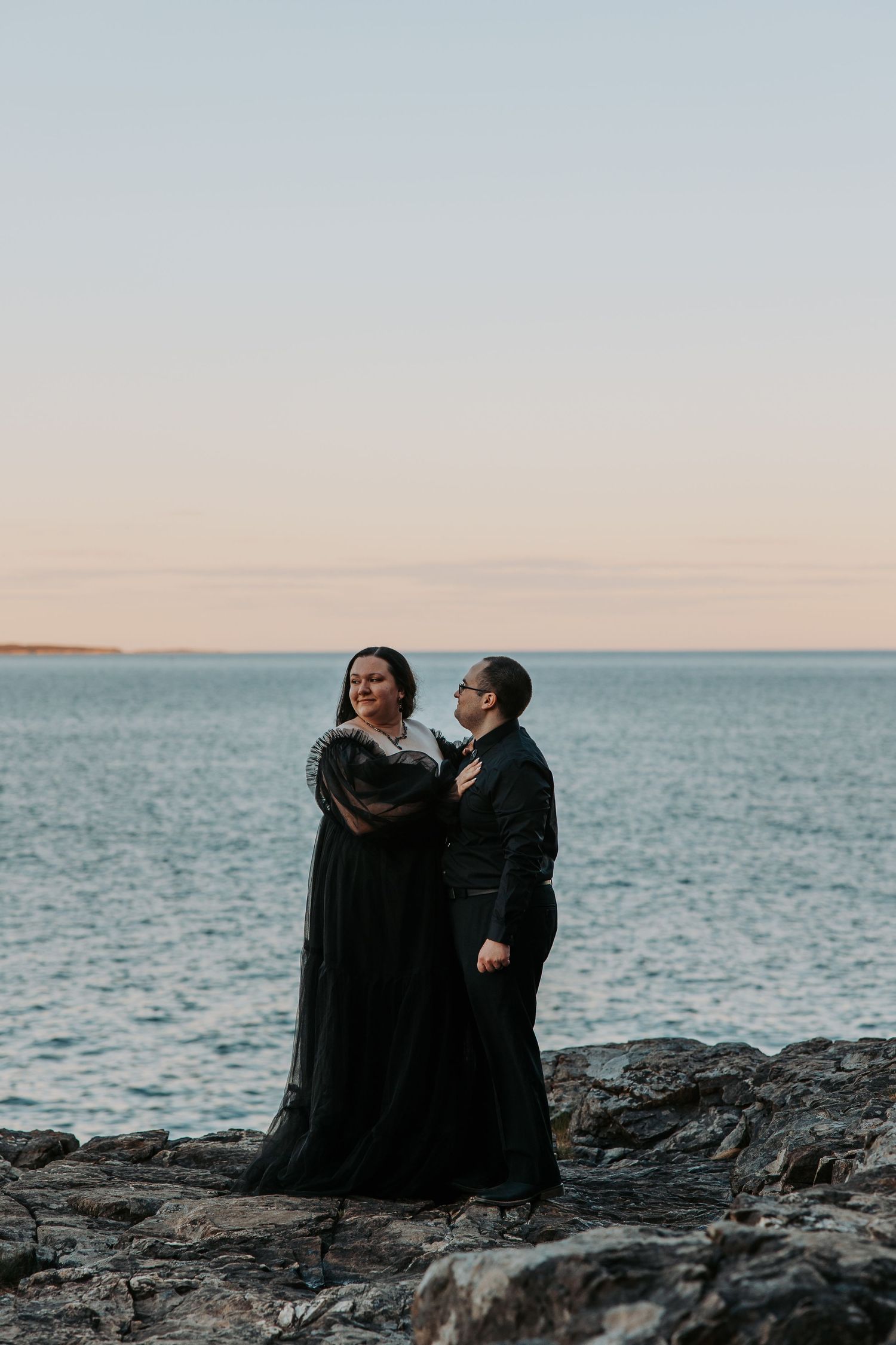 Two figures in dark clothing sit together on coastal rocks overlooking a serene ocean view at dusk.