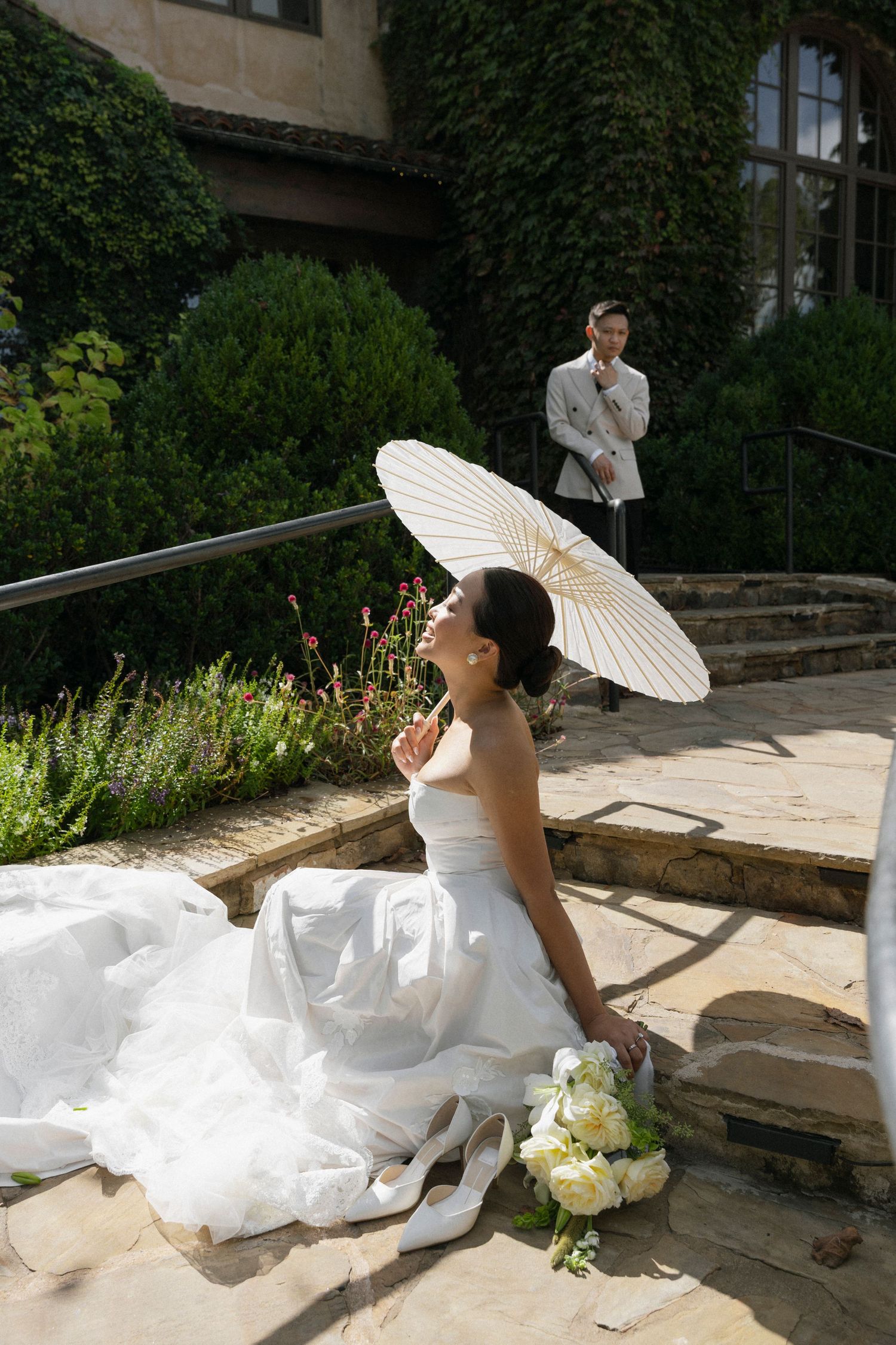 A bride in a flowing white dress poses with a decorative white parasol on stone steps surrounded by lush greenery.