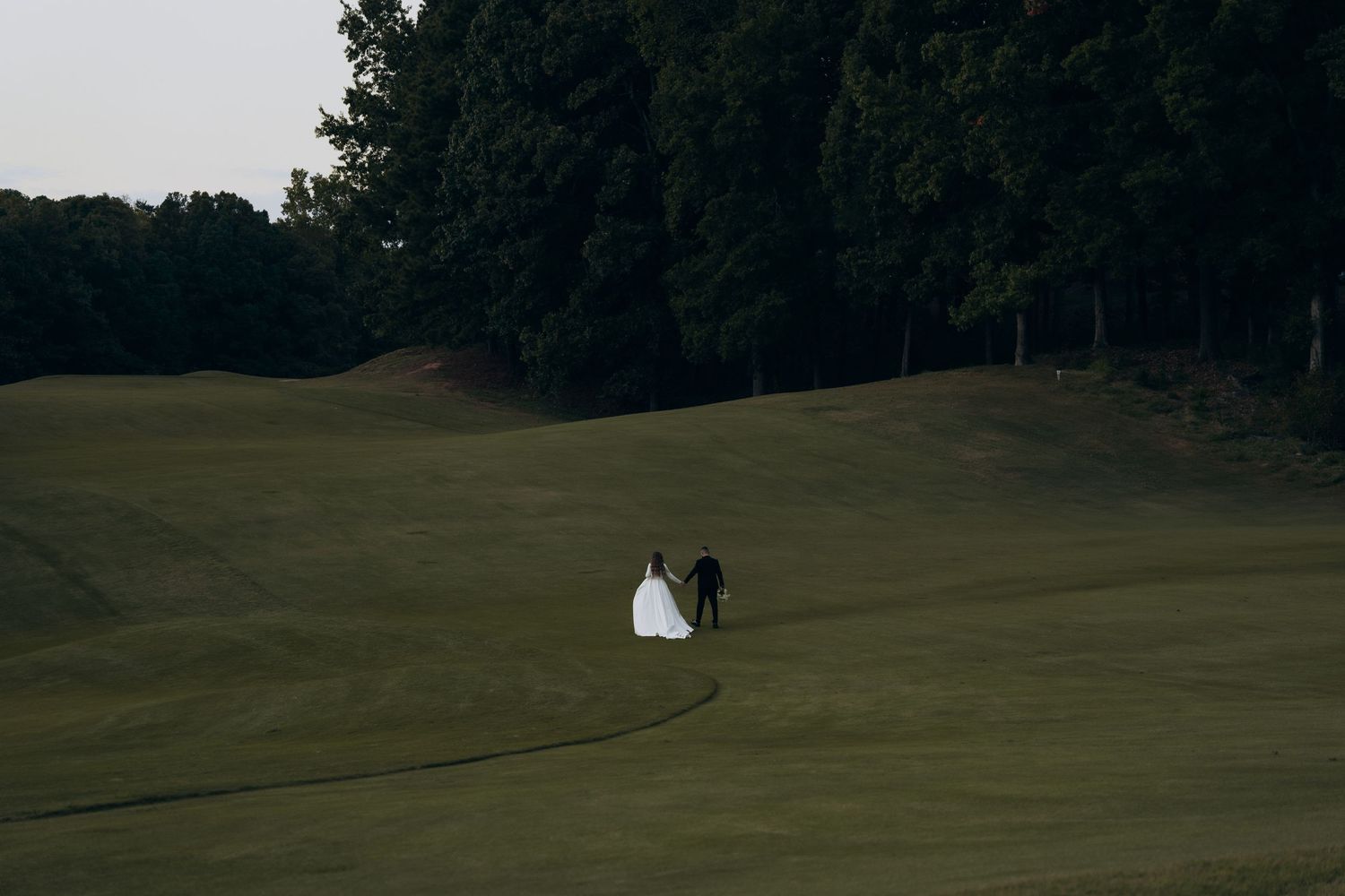 A couple in wedding attire walks across a lush green golf course fairway bordered by trees.