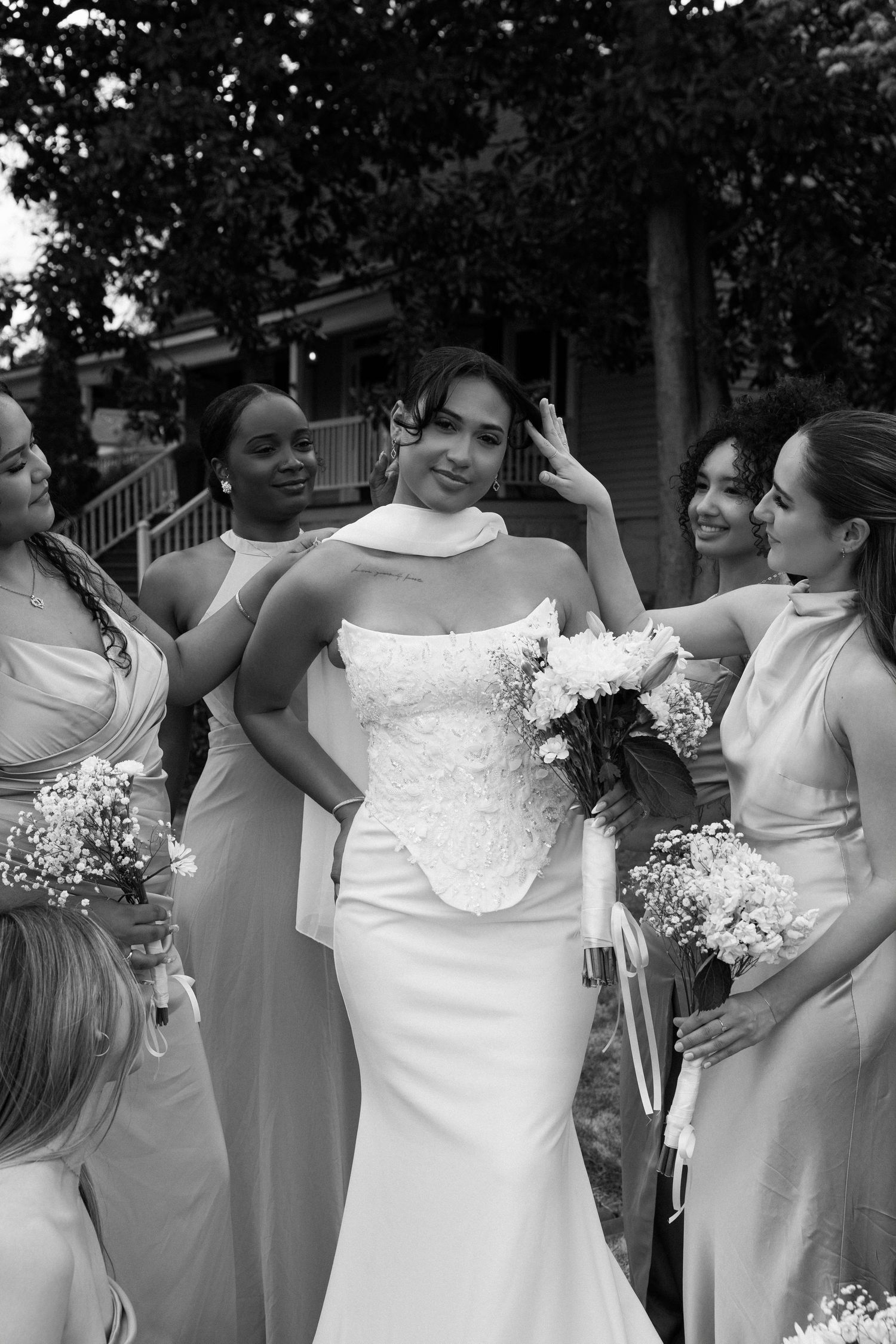 A bride in a white strapless gown poses with bridesmaids in light-colored dresses holding bouquets during an outdoor wedding.