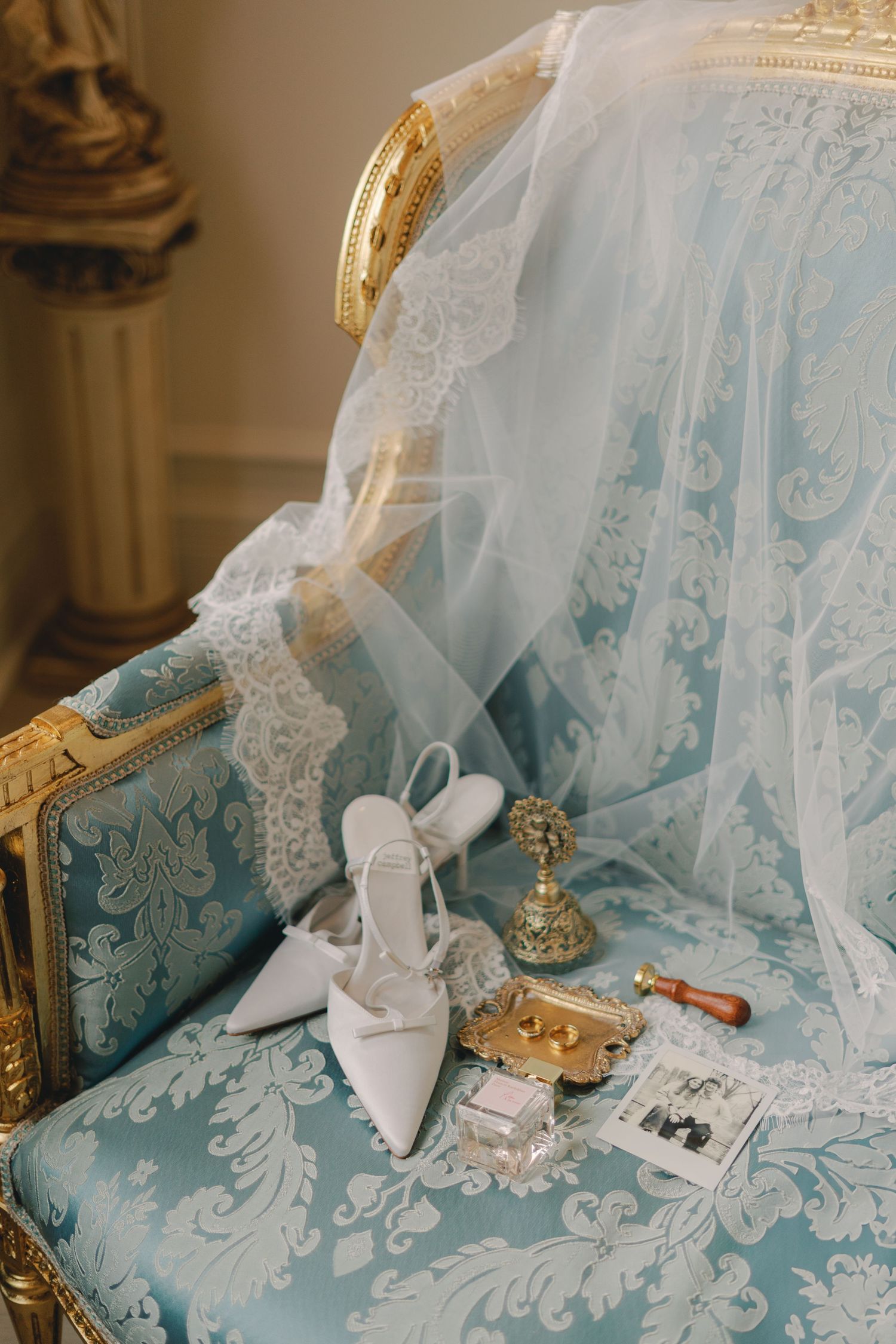 White bridal heels and wedding accessories displayed on an ornate blue damask chair with lace veil draped over it.