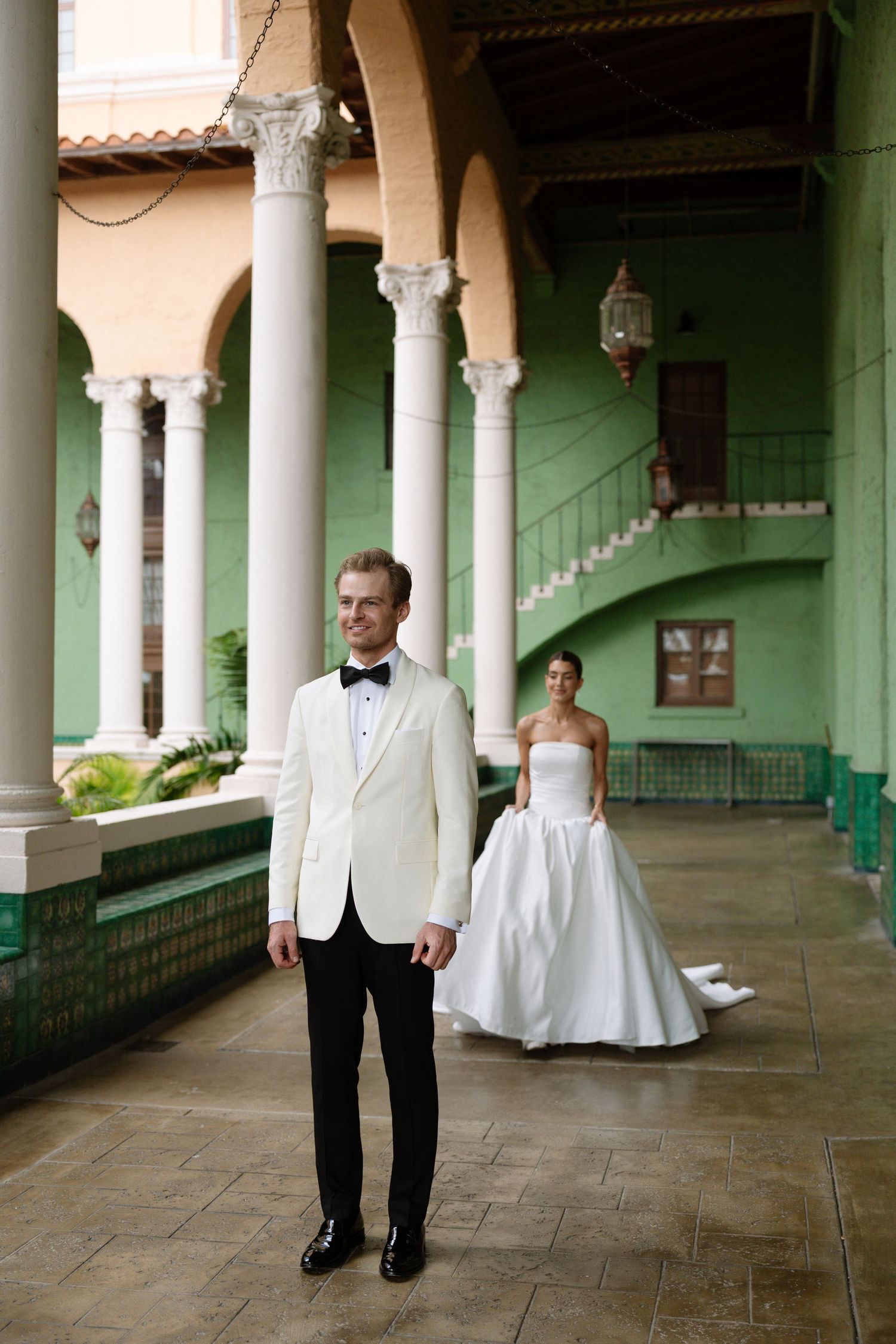 Groom in white tuxedo jacket stands in green courtyard colonnade as bride in strapless gown approaches behind him.