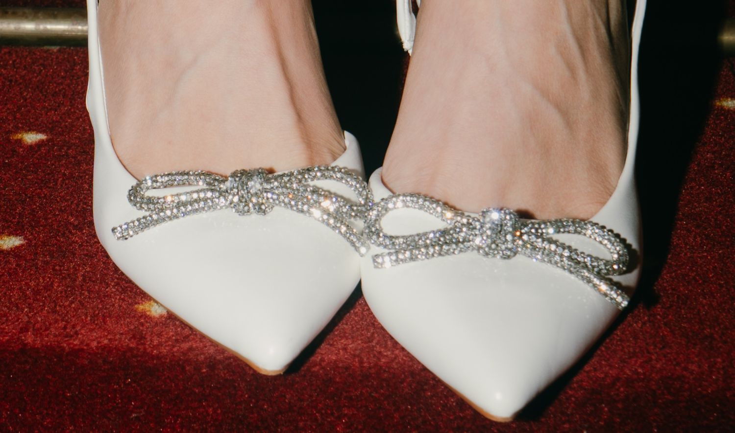Elegant white bridal shoes featuring sparkly crystal bow designs on burgundy carpet.