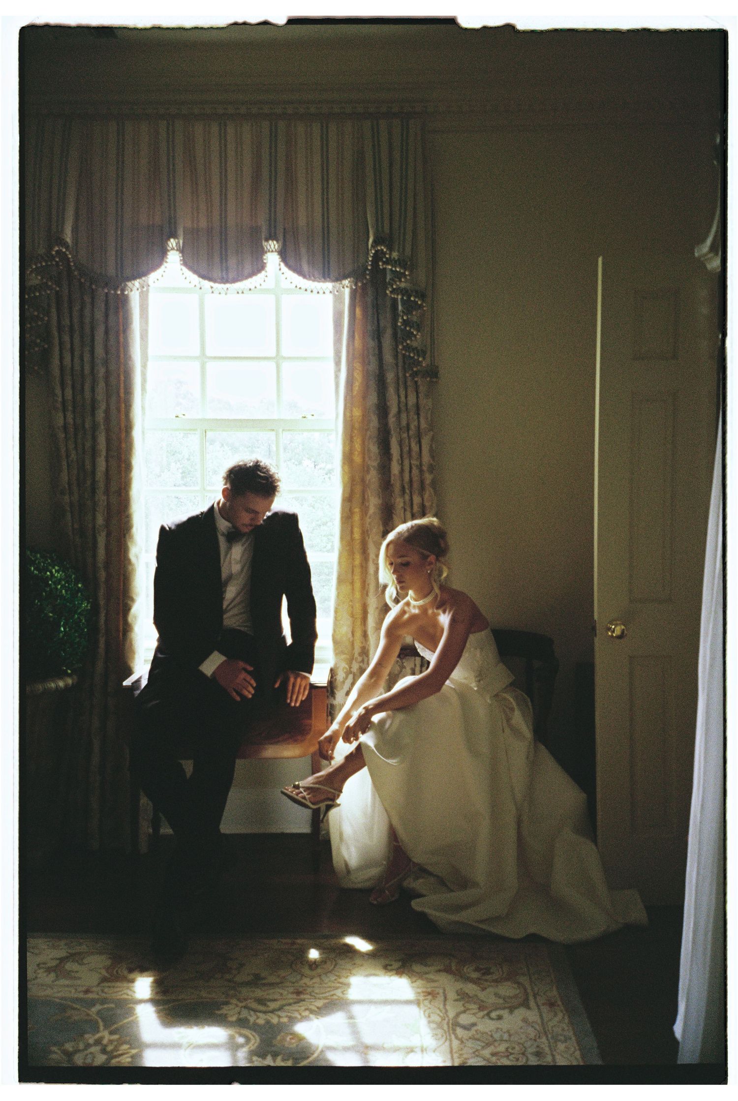 Bride adjusting her heels while groom sits nearby by a sunlit window in an elegant room on their wedding day.