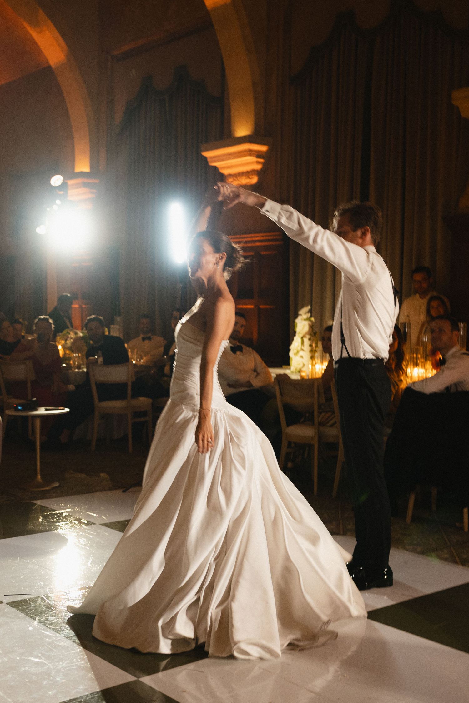 Bride and groom share first dance on checkered floor in grand ballroom with guests watching in warm light.