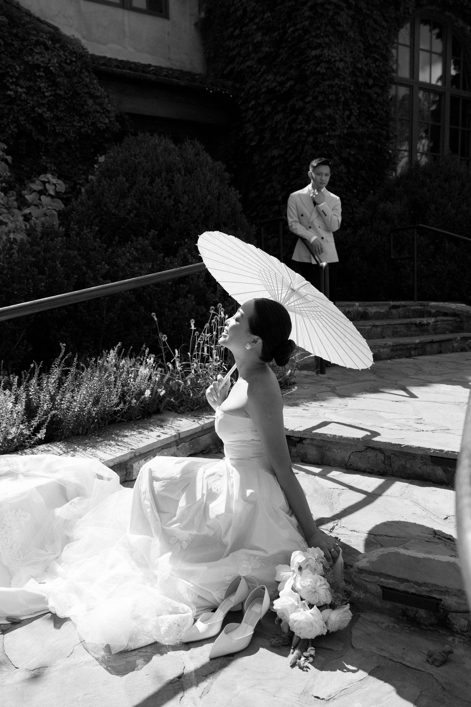 A black and white wedding photo featuring a bride in a flowing dress holding a white parasol on stone steps.