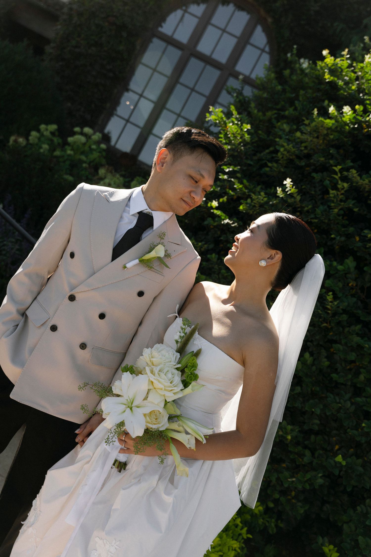 A romantic wedding couple in white attire embraces in a garden setting with white lilies and greenery.