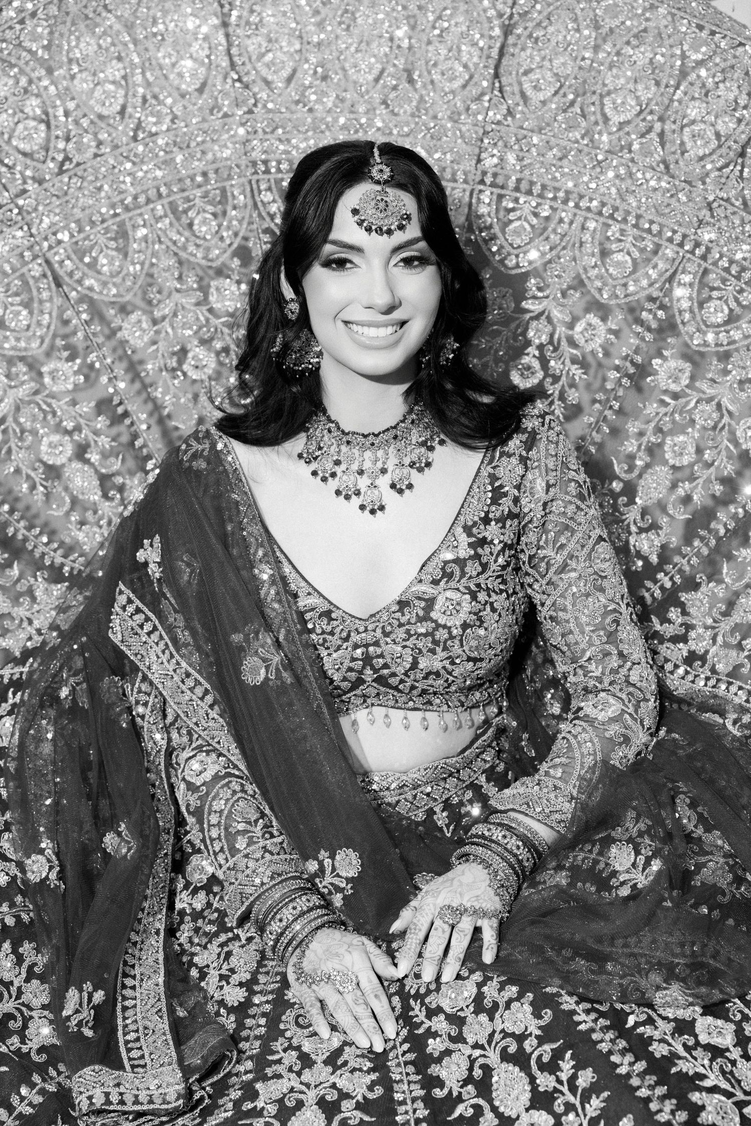 A woman in traditional Indian bridal attire with ornate jewelry and embroidered clothing poses against a decorative backdrop.