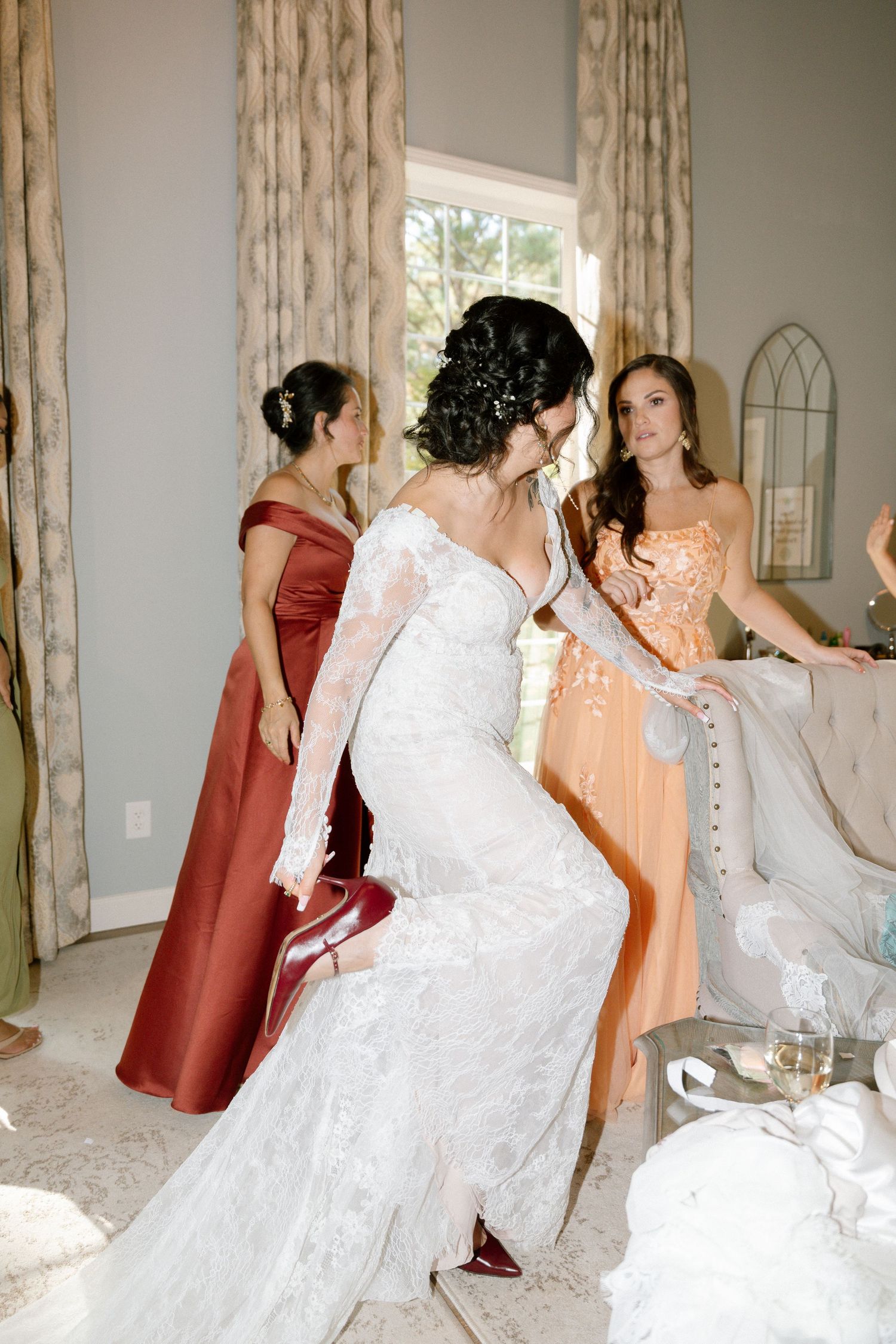 Bridesmaids in burgundy dresses help adjust a bride's white lace wedding gown before the ceremony.