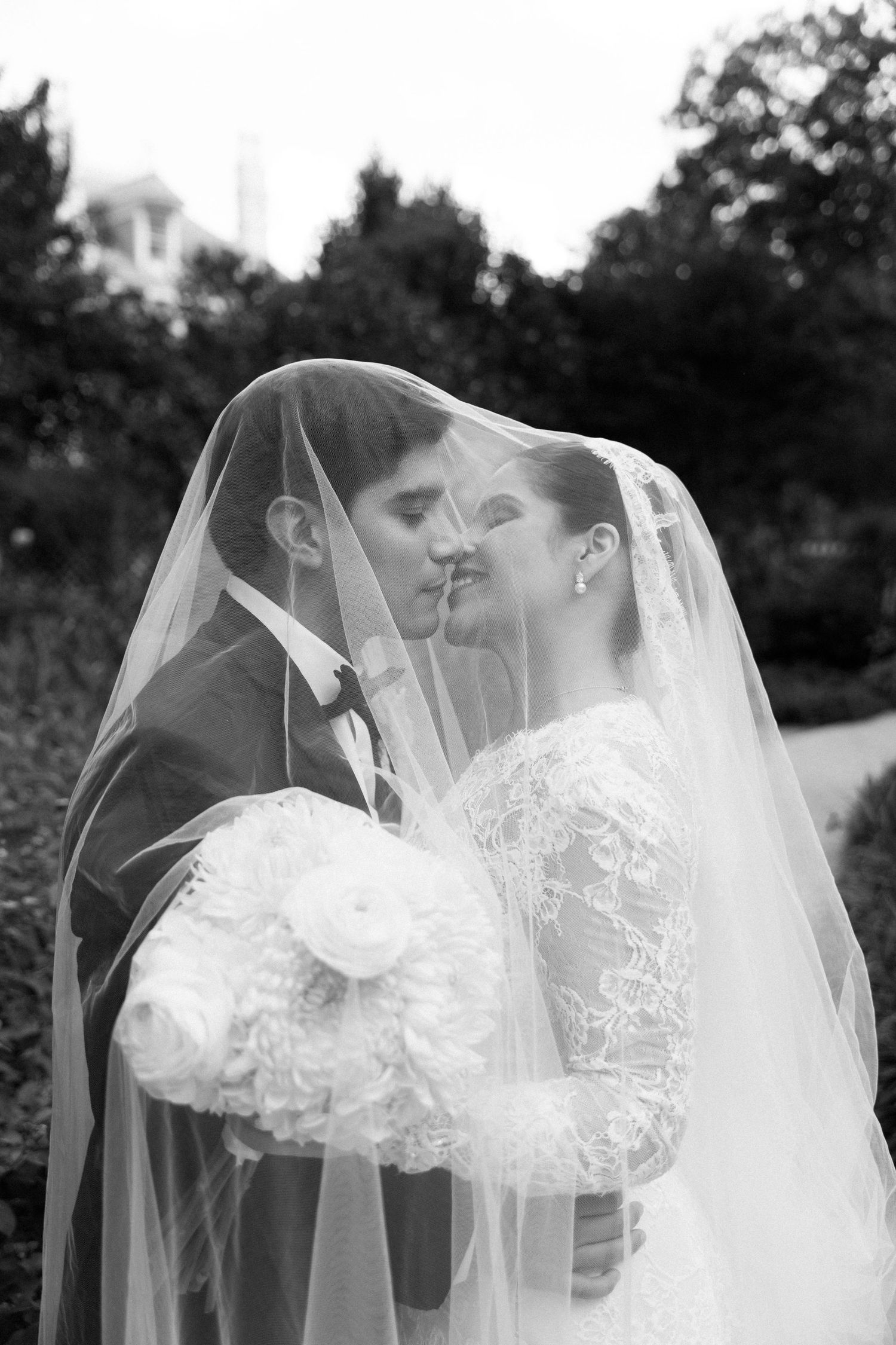 A romantic black and white wedding photograph showing a tender moment under a delicate veil in an outdoor garden setting.