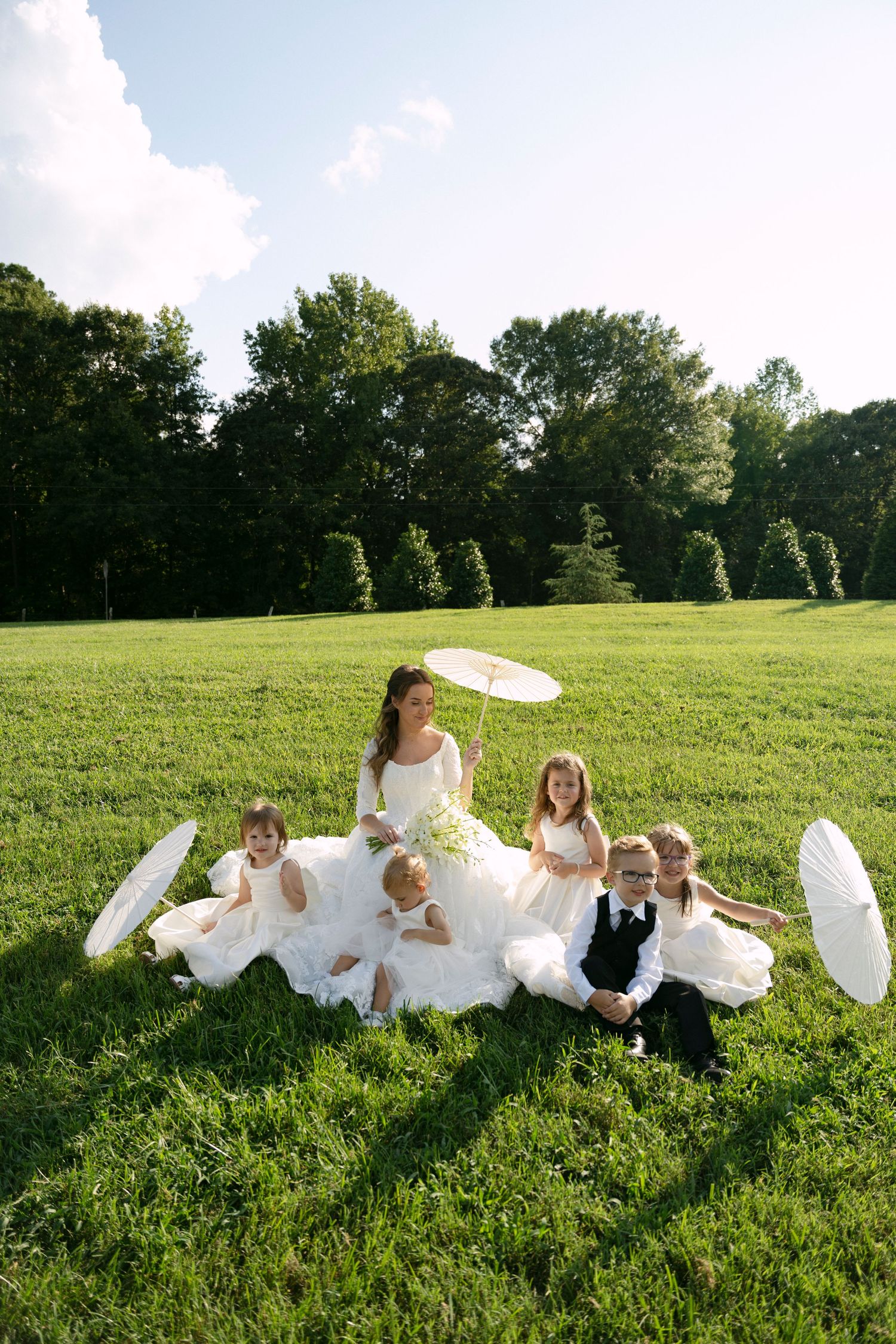 A group in formal attire lies playfully in a green meadow on a sunny day, creating a whimsical scene with white fabric.