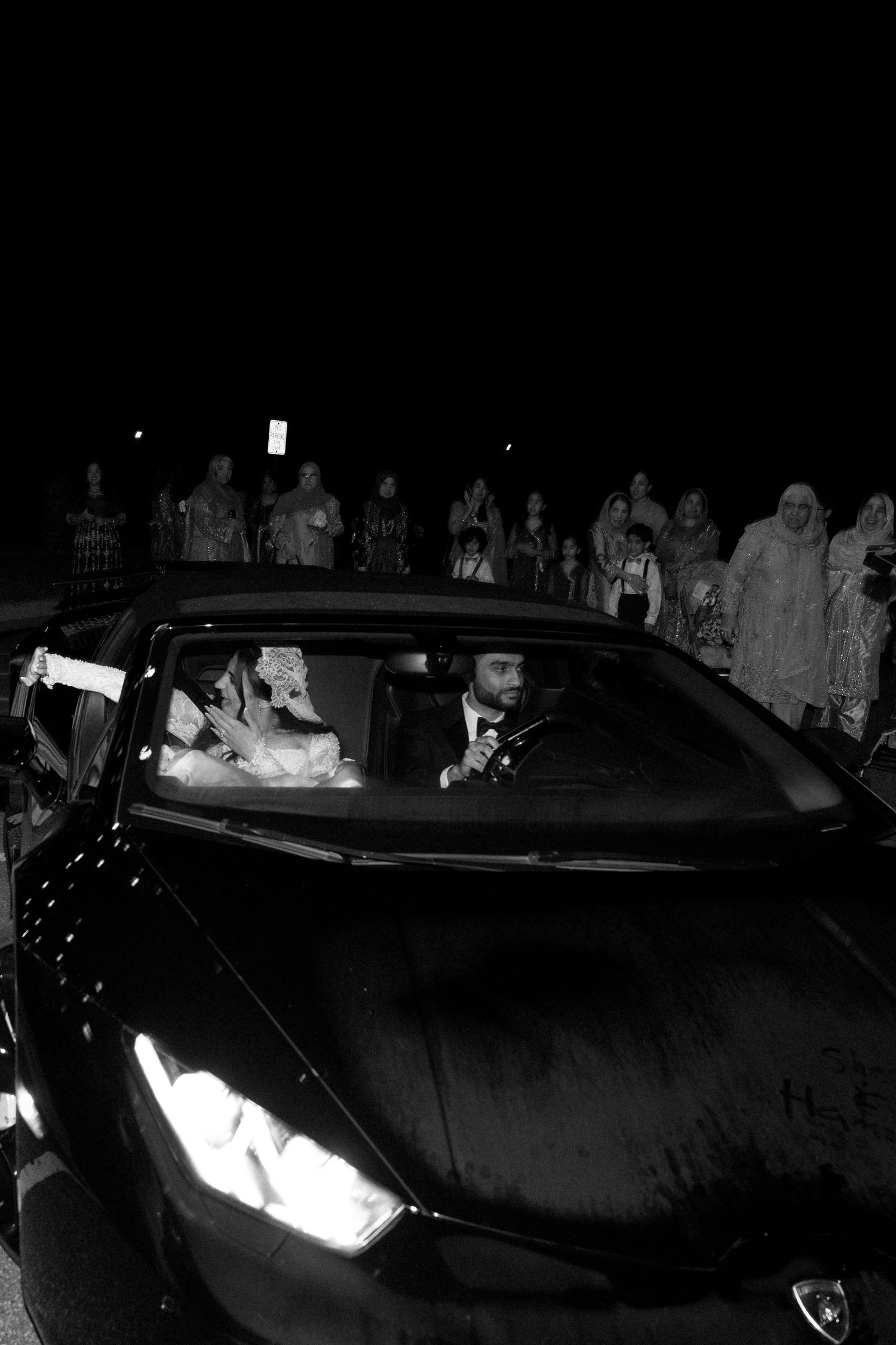 A dark nighttime scene shows car headlights illuminating a group of figures standing in the shadows.