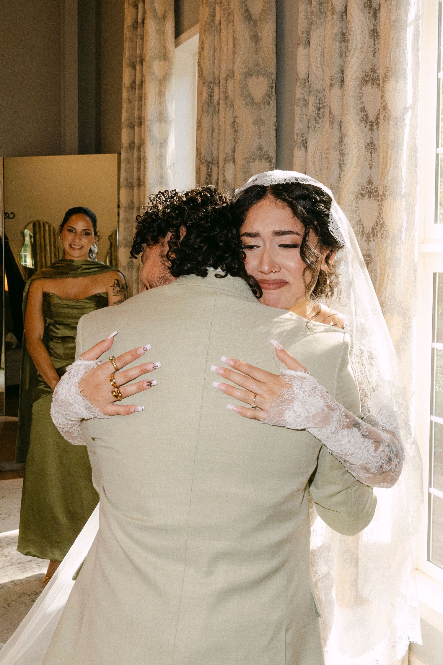 A bride in a white lace dress embraces someone in a beige suit during an emotional wedding moment near sheer curtains.