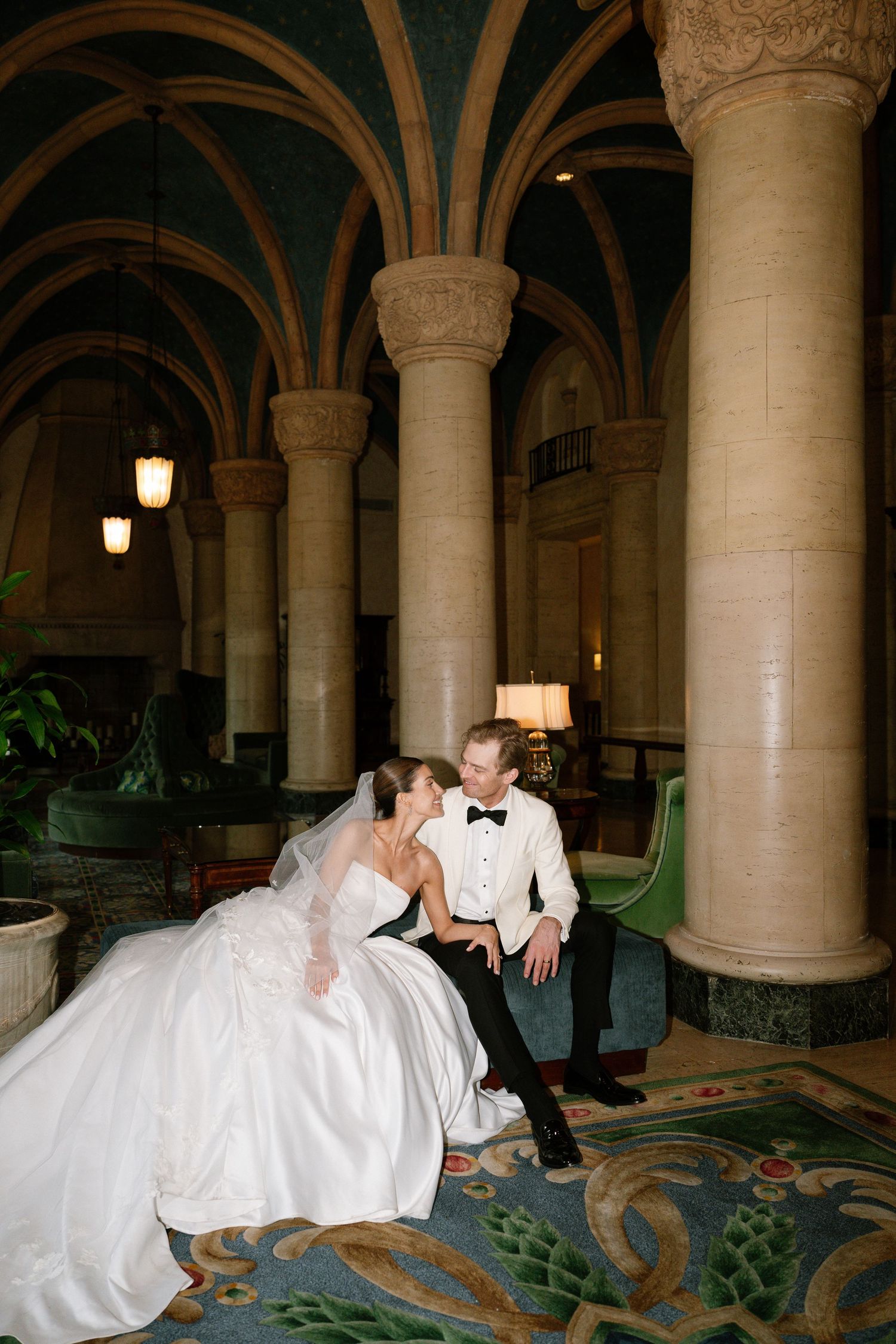 Bride and groom share an intimate moment seated among grand arched columns in an elegant hotel lobby.