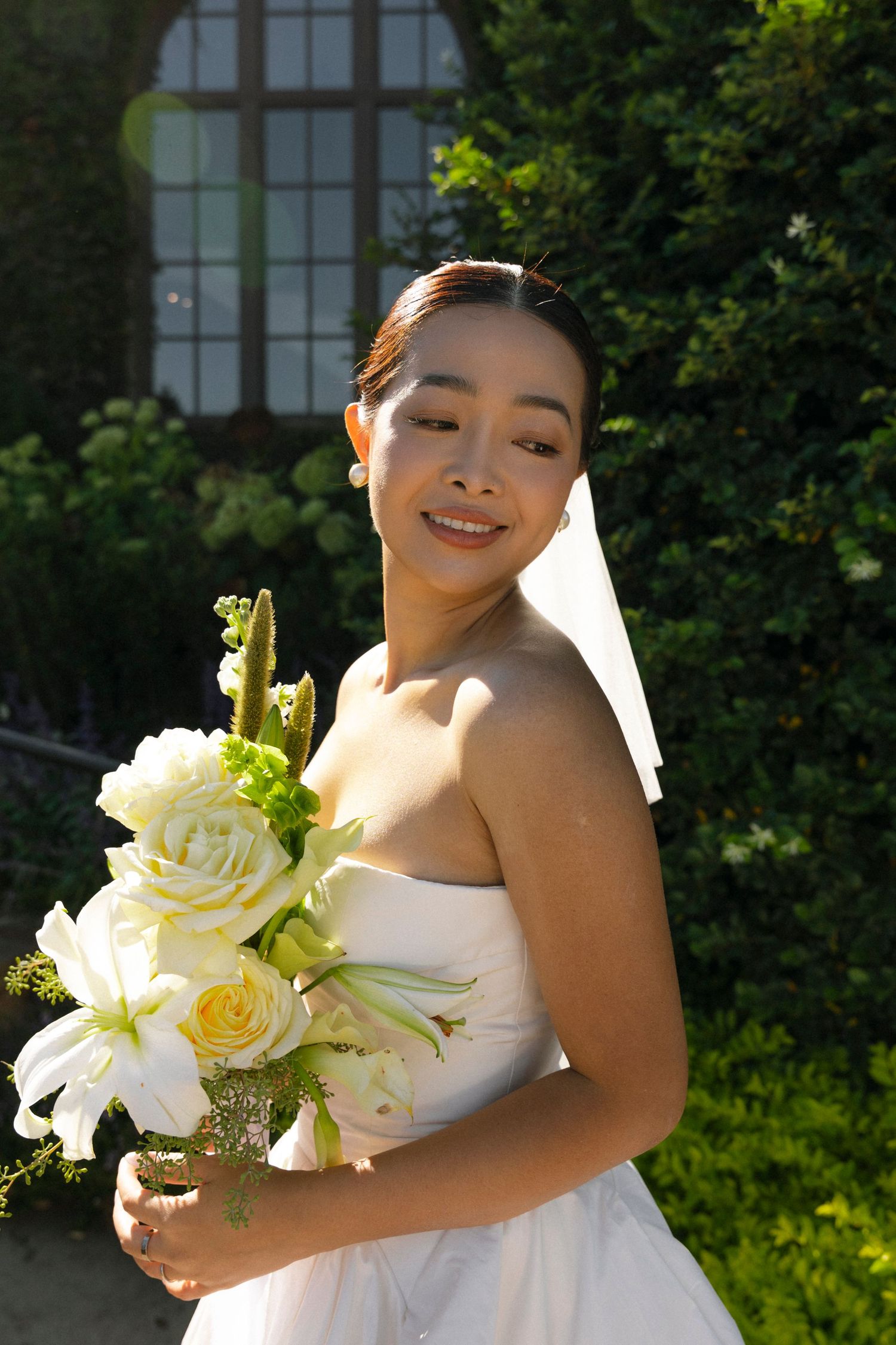 A bride in a white strapless wedding dress holds a beautiful bouquet of white roses and lilies in a sunny garden setting.