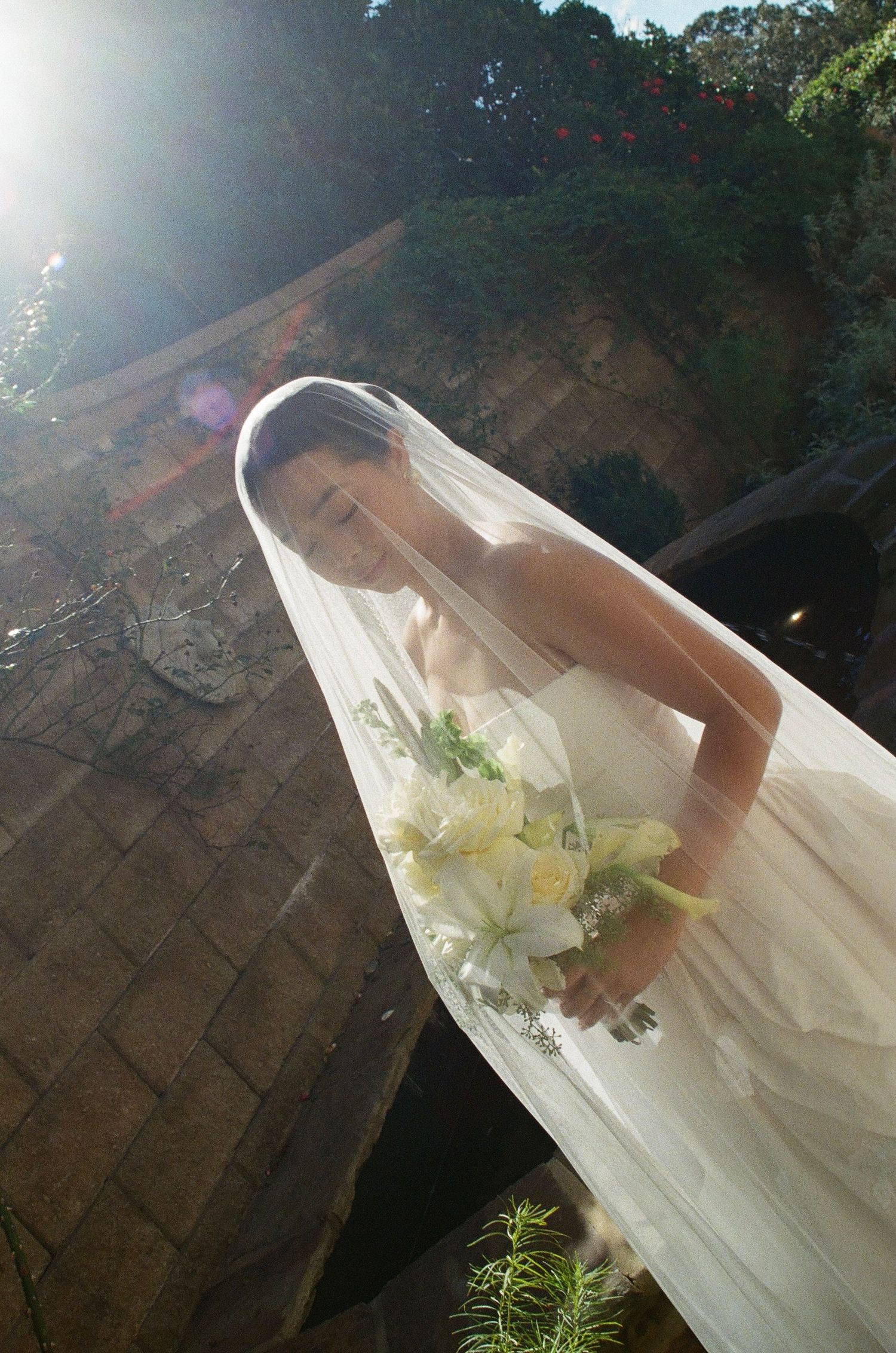 A bride in a flowing white veil and dress holds a bouquet of white lilies while standing in dappled sunlight outdoors.