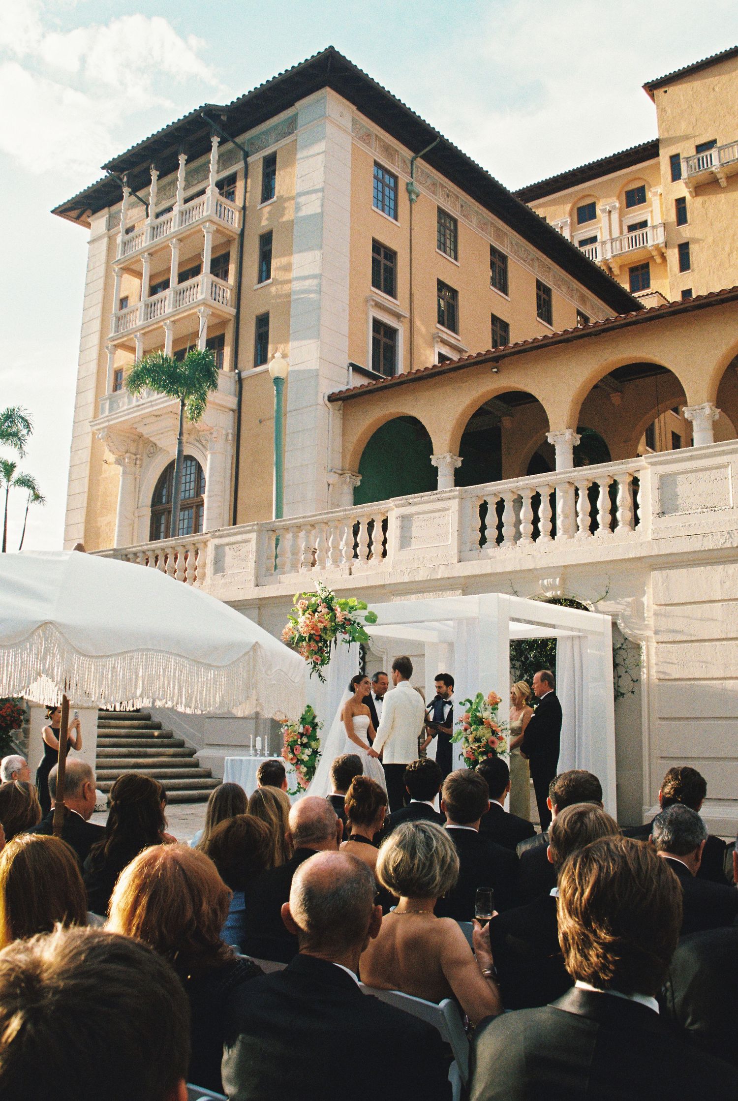 Outdoor wedding ceremony at elegant Mediterranean-style venue with guests seated before couple at floral altar.