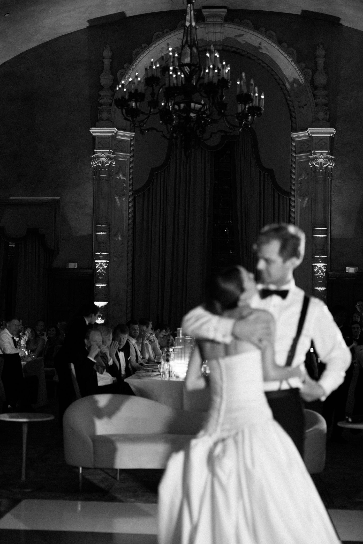 Bride and groom share their first dance in an elegant ballroom with chandelier and arch in black and white photo.