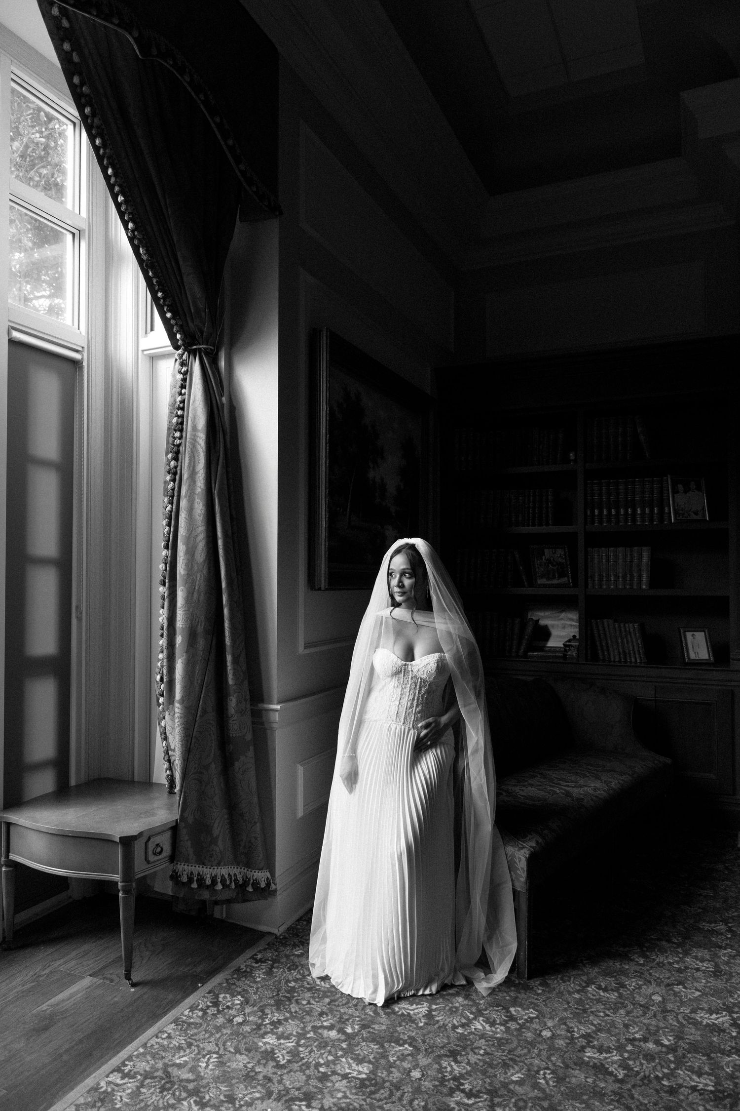 A ghostly figure in a white dress and veil stands in a dark Victorian-style room beside a window.