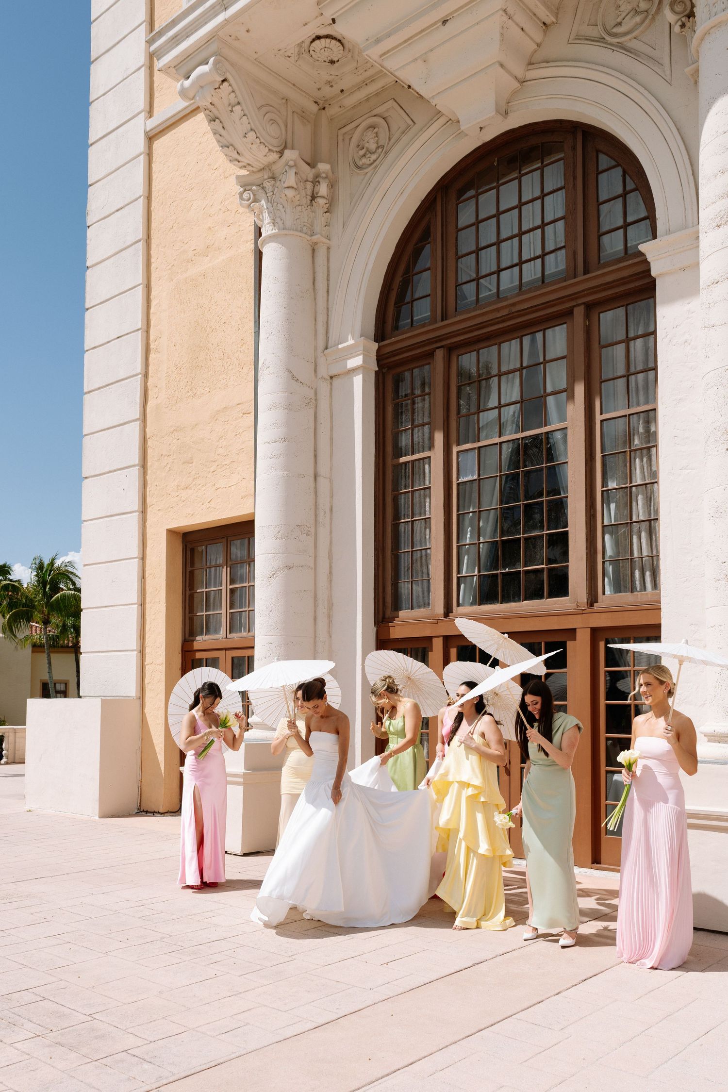 Bride and bridesmaids in pastel gowns holding white parasols outside an elegant building on a sunny wedding day.