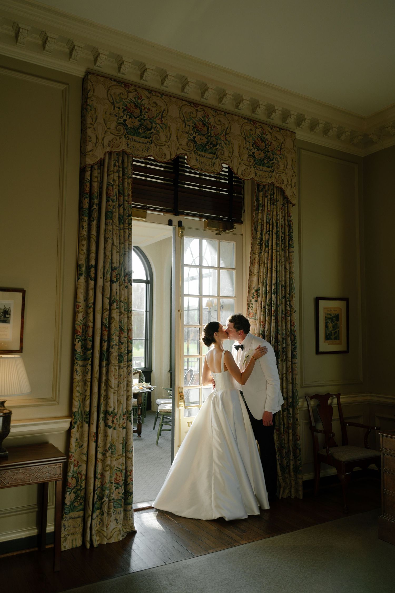 Bride and groom share a romantic kiss in an elegant room with floral drapes and arched windows in a historic venue.