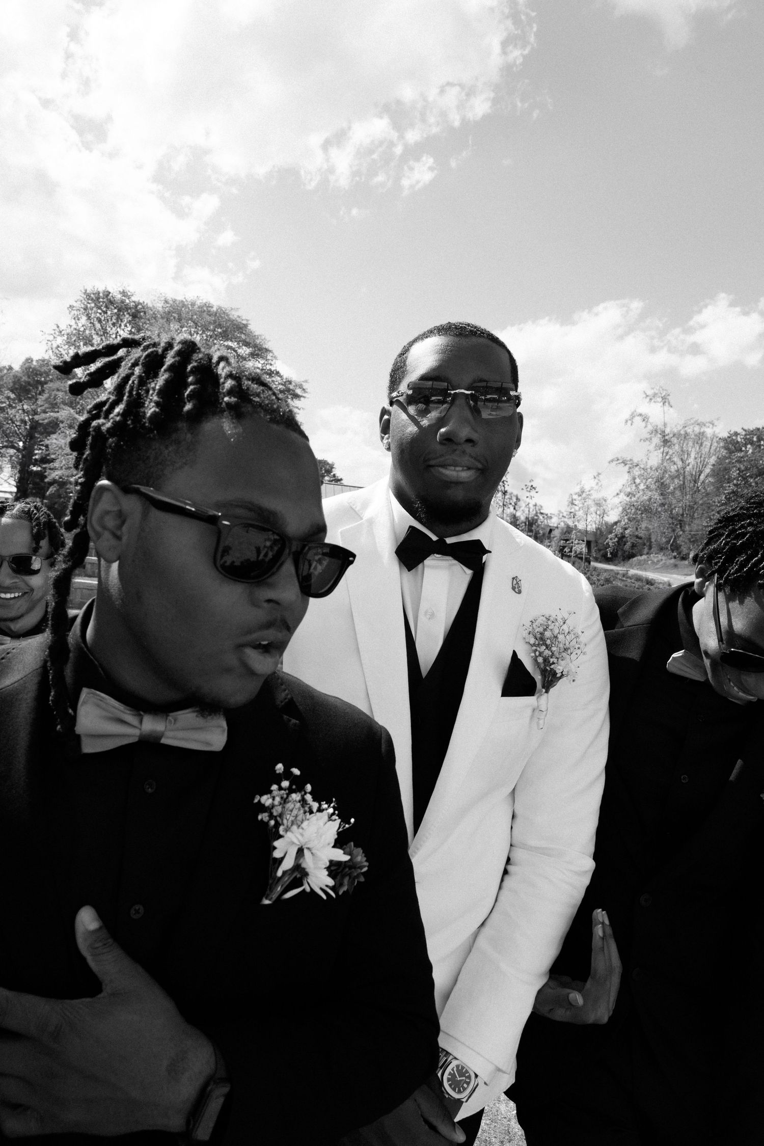 Black and white photo of wedding guests in formal attire with bow ties and sunglasses at an outdoor celebration.