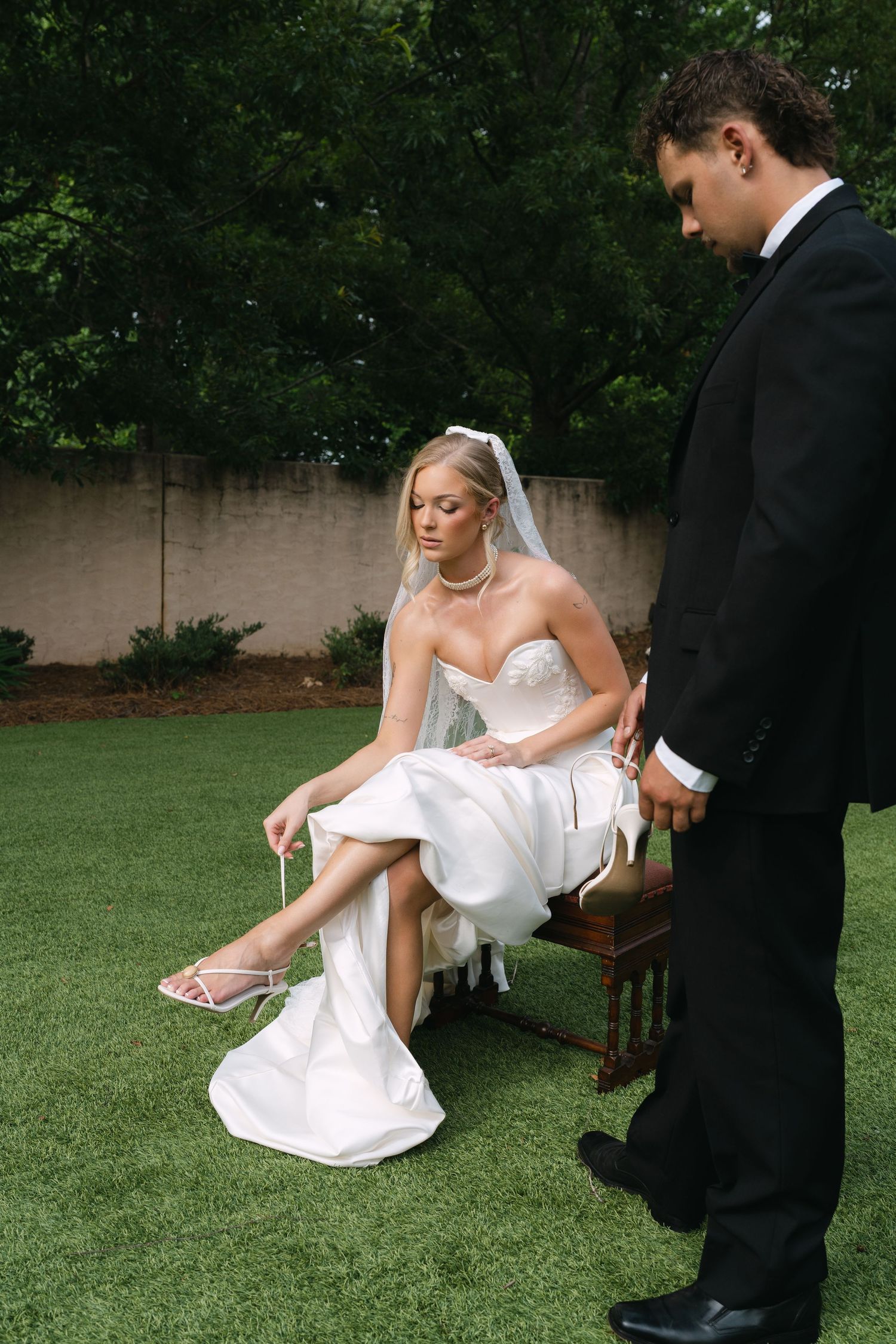 Groom in black tuxedo helps bride in white strapless gown adjust her heels while seated outdoors on wedding day.