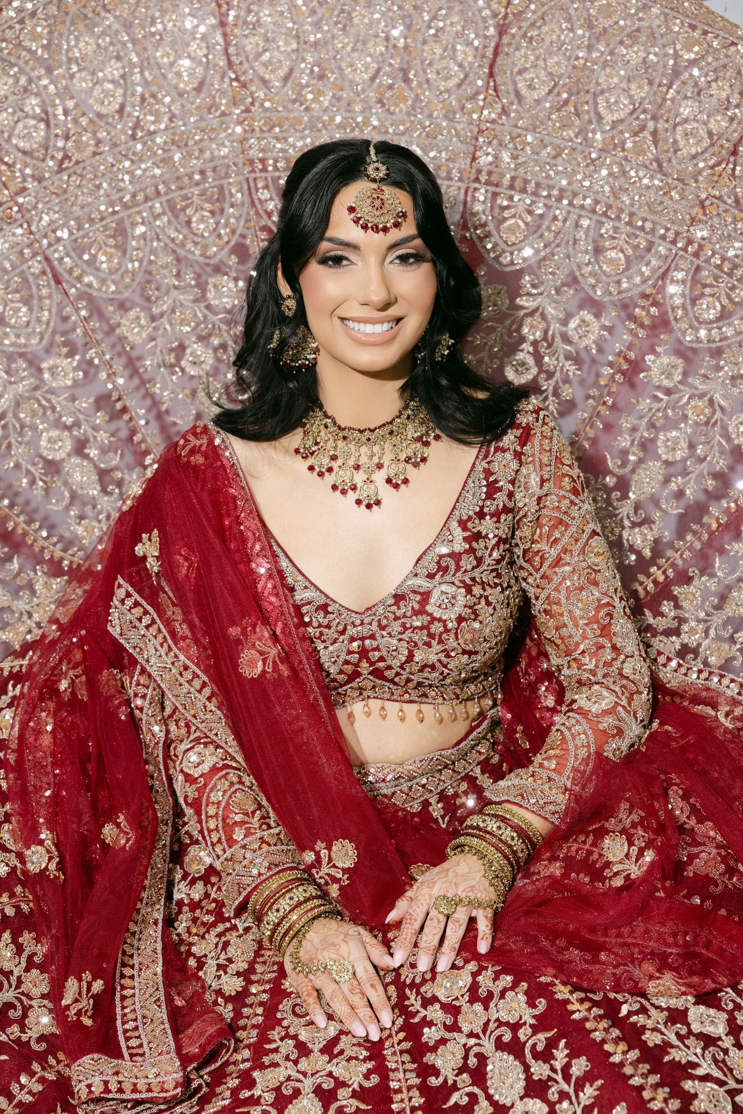 A bride wearing an ornate red and gold embroidered Indian lehenga poses against a sparkly backdrop.