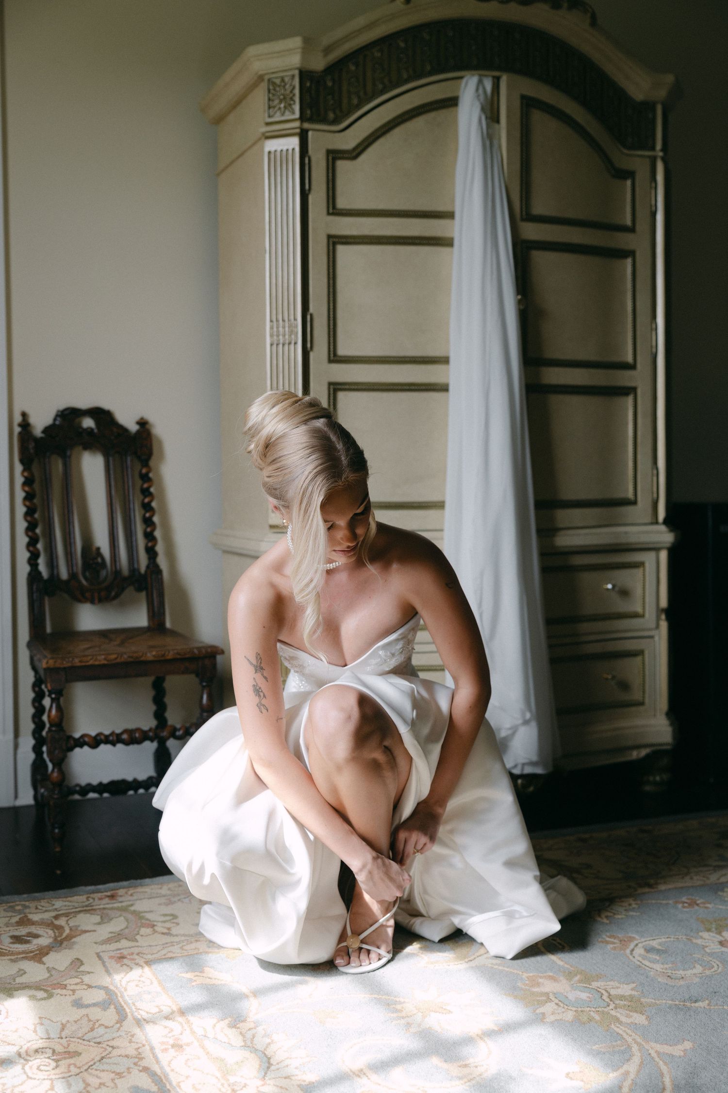 Bride in white strapless gown sitting on floor fastening her heels in an elegant vintage room.