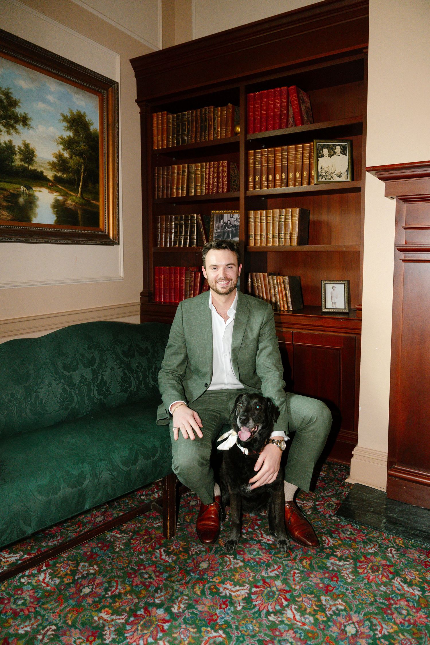 A person in a green suit sits with a black dog on an emerald velvet couch in a study with bookshelves and artwork.