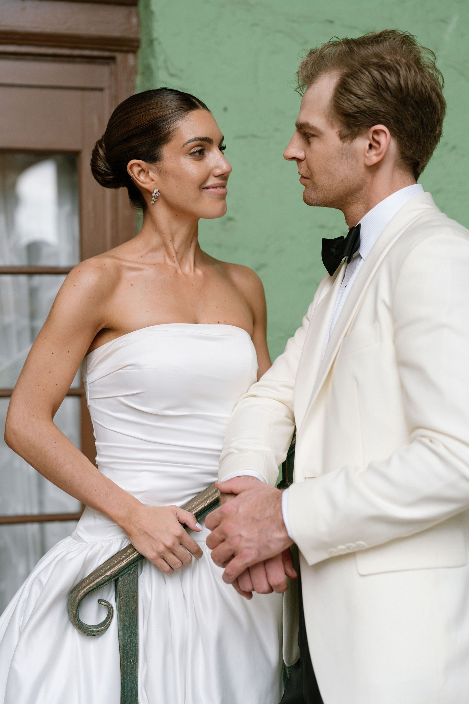 Bride in strapless white gown and groom in white tuxedo share an intimate moment outdoors against a green wall.