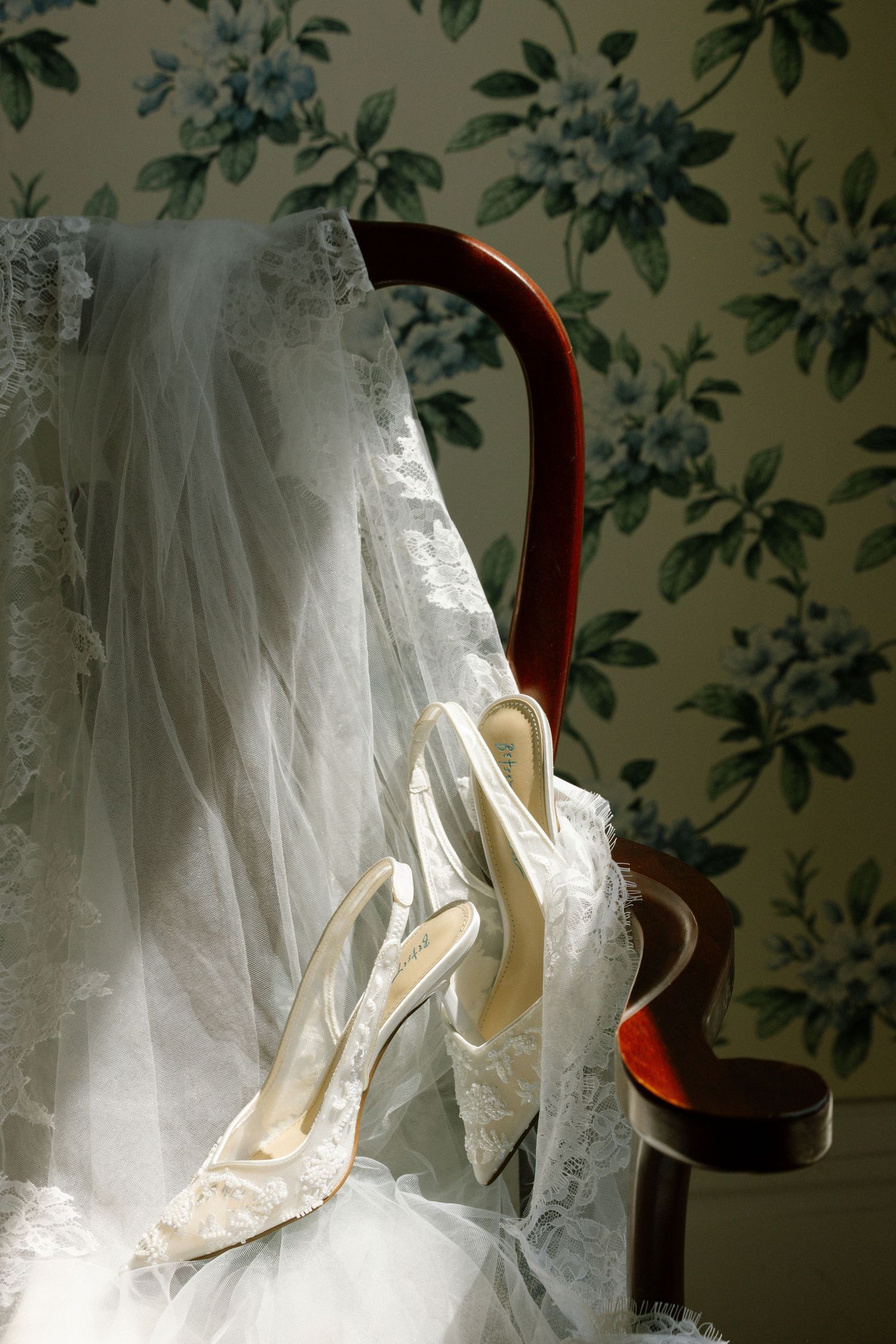 Elegant white wedding dress and ivory bridal heels arranged on wooden chair against floral wallpaper background.