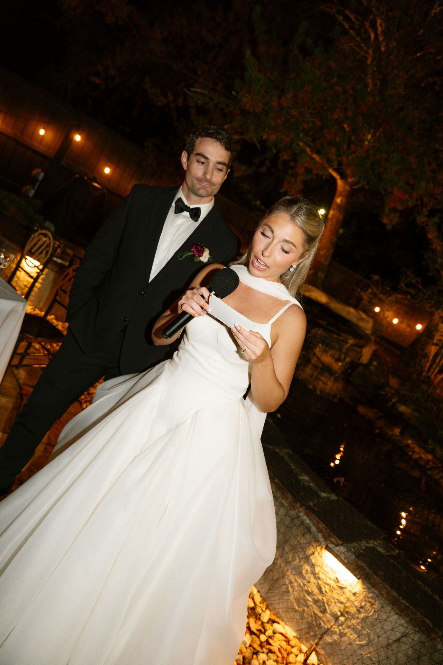 A couple in formal wedding attire stands outdoors at night with ambient lighting and autumn leaves on the ground.