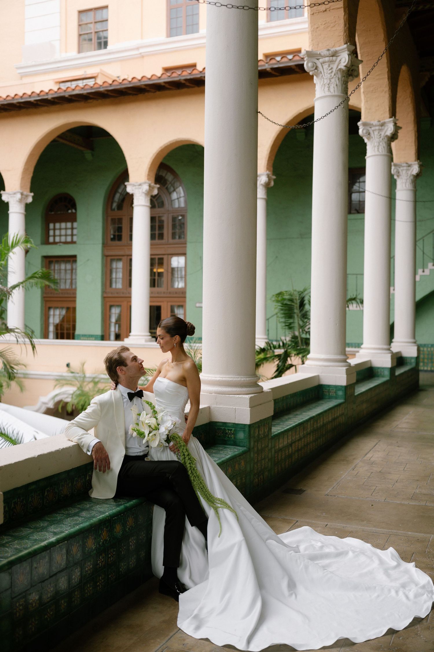 Bride in white strapless gown and groom in tuxedo pose by elegant arched columns at a classic venue.