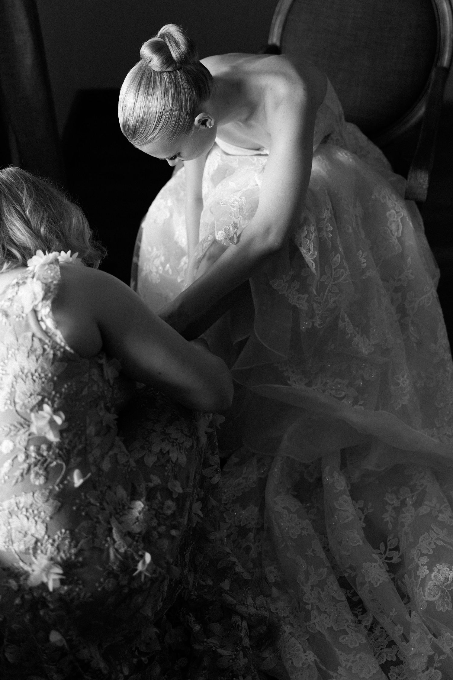 Black and white photo of a bride in lace gown being helped by bridesmaid while seated in elegant chair.