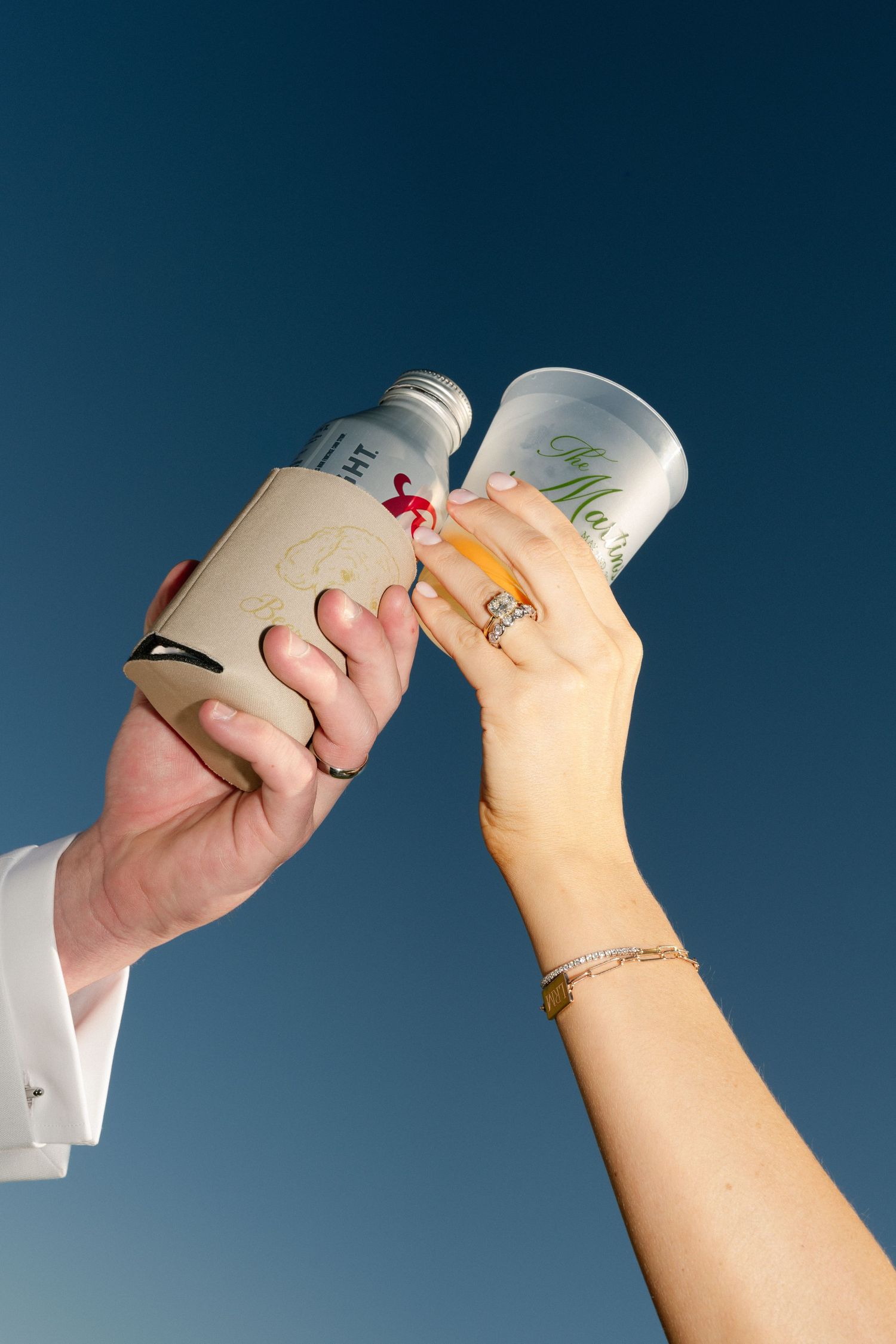 Two hands toasting drinks against a blue sky, one wearing a diamond ring and gold bracelet.