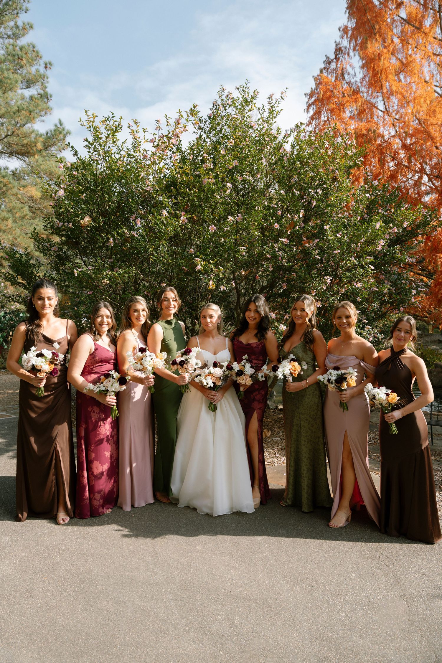 Wedding party in autumn with bridesmaids wearing mix of brown and pink dresses holding floral bouquets outdoors.