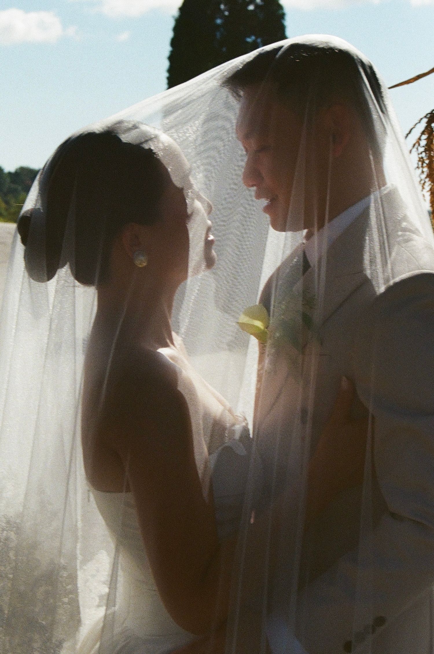 A romantic silhouetted wedding couple shares an intimate moment behind a sheer white veil in soft lighting.