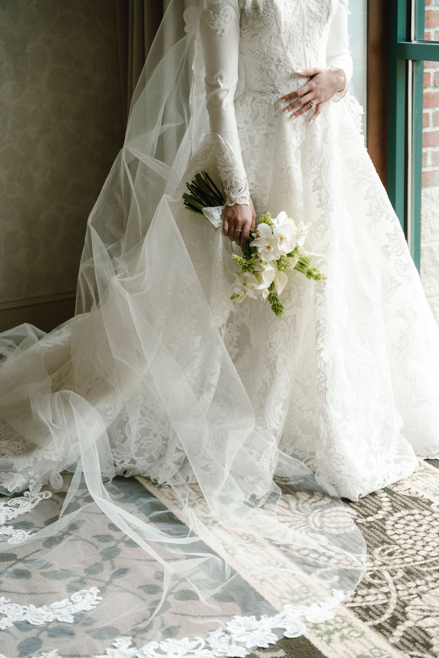 Close-up detail of a flowing white lace wedding dress and veil with delicate floral bouquet against a window.