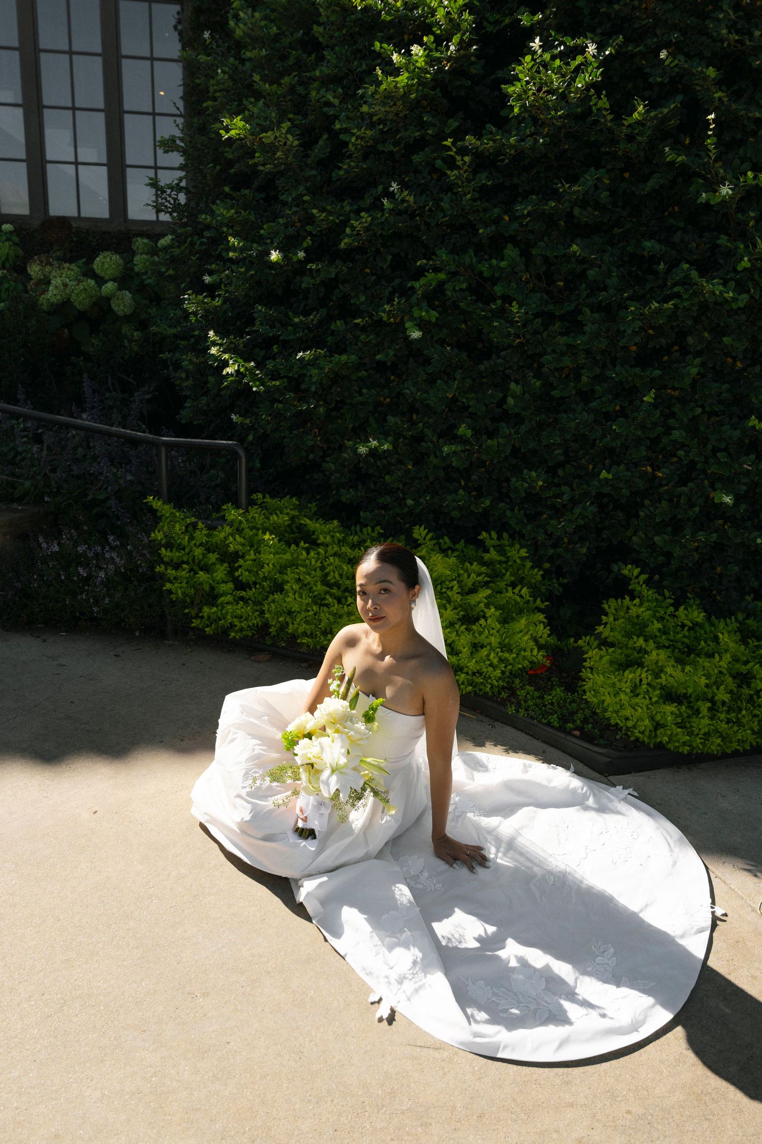 A bride in a white strapless gown sits gracefully on the ground holding a bouquet of white flowers in a garden setting.