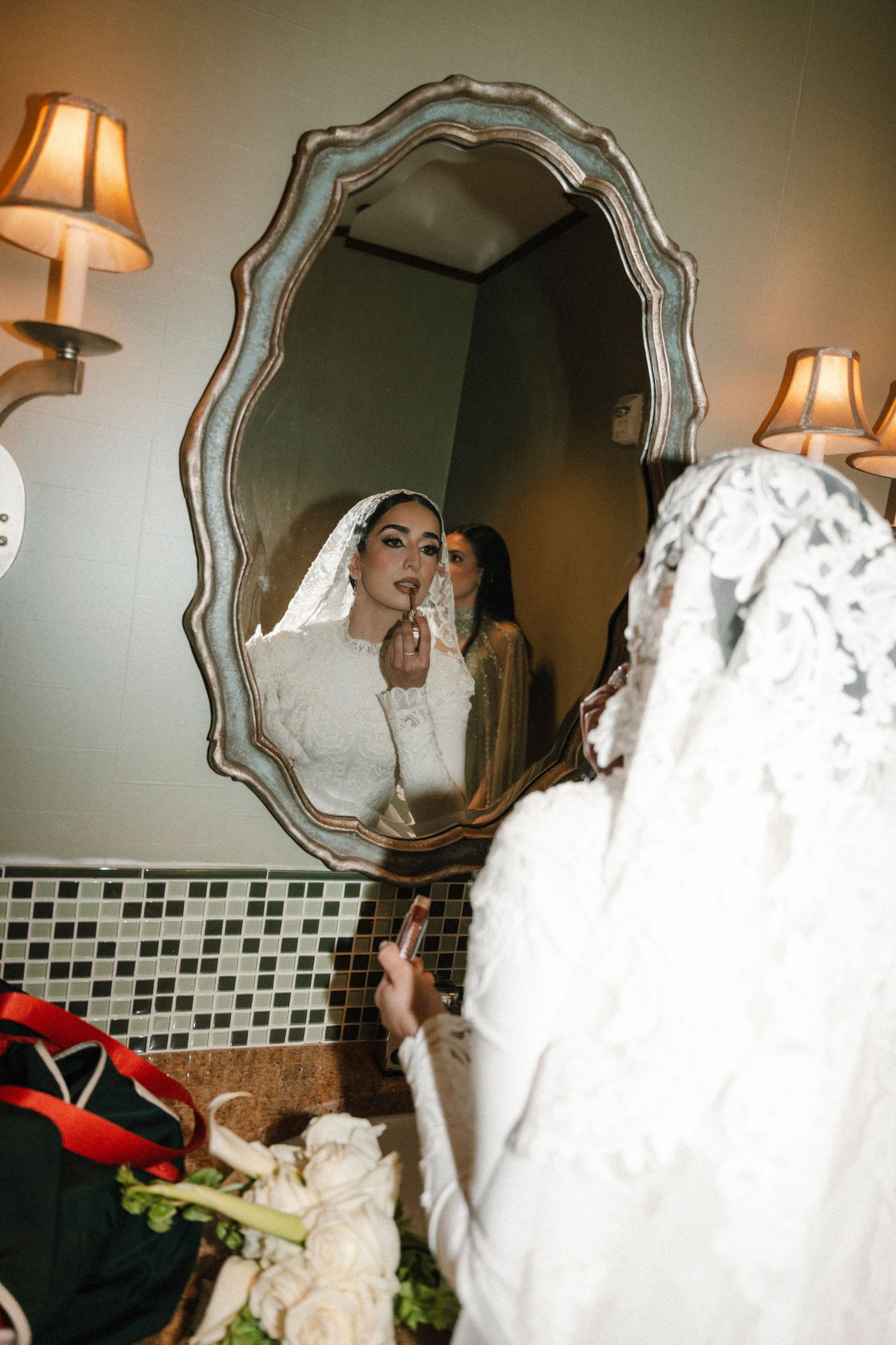 A reflection in an ornate mirror shows someone in a white wedding dress getting ready with warm lighting and wall sconces.