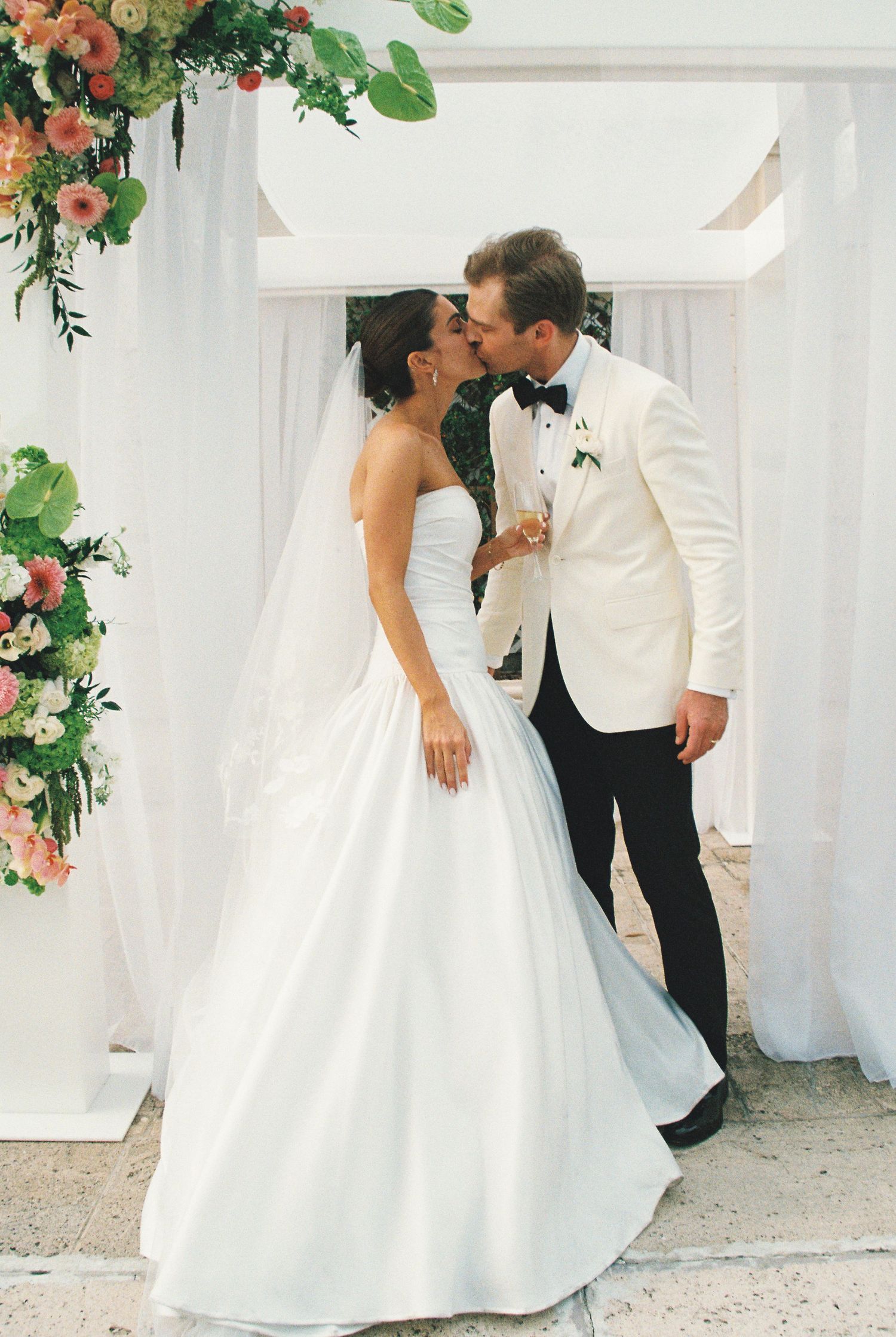 Bride and groom sharing a kiss at outdoor wedding ceremony, white draping and floral arch backdrop.