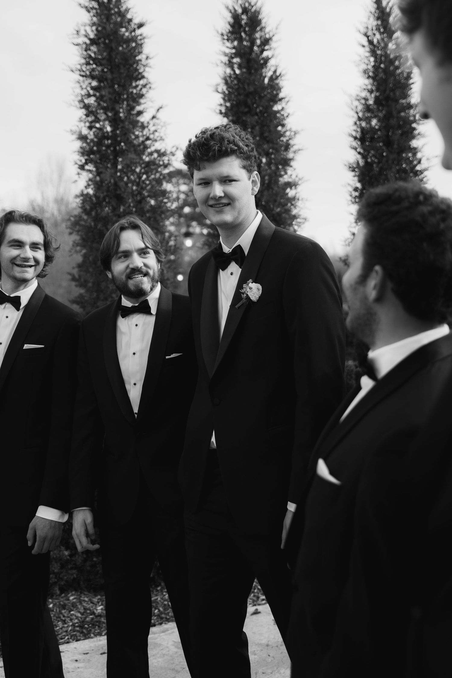 A black and white photo of a wedding party sharing a moment outdoors with evergreen trees in the background.