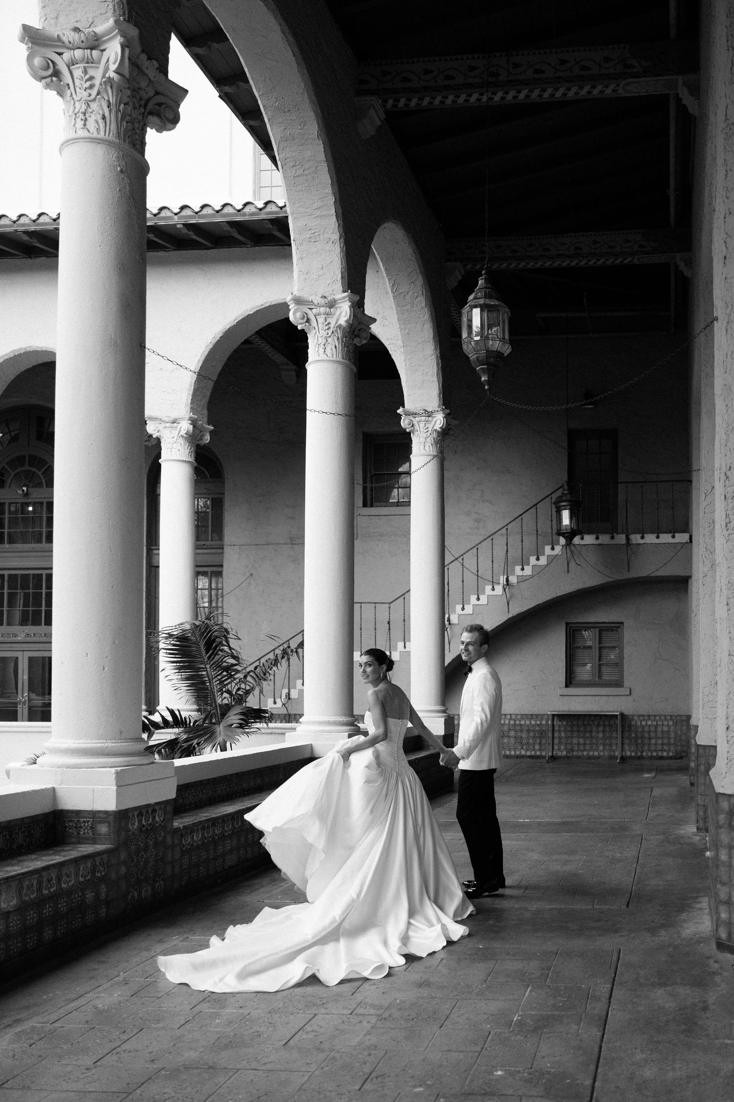 Black and white photo of bride and groom holding hands under elegant arched colonnade at a classic wedding venue.