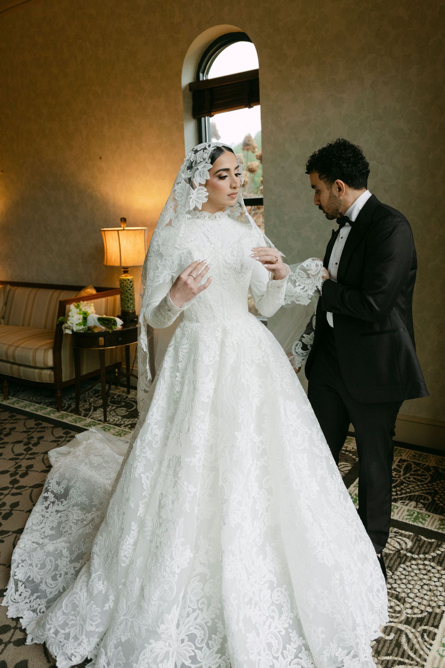 A bride in an elegant white lace gown and groom in a black suit share intimate moments during their wedding photoshoot.