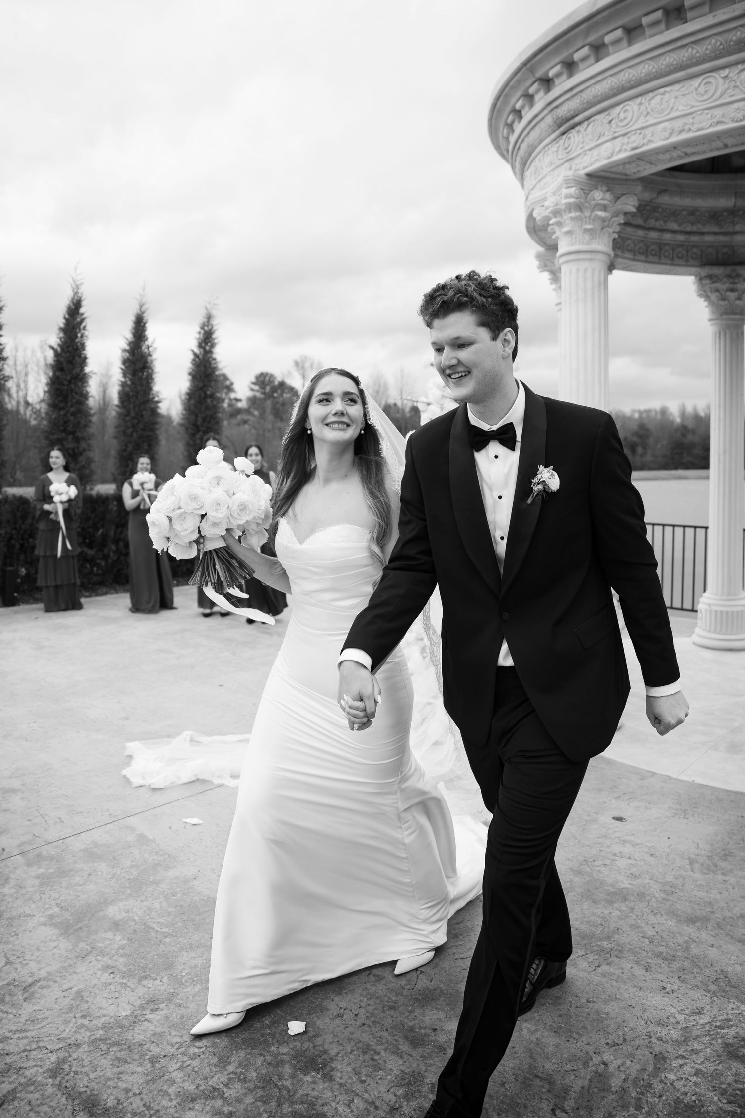A happy newlywed couple walks hand in hand near a columned gazebo in black and white photography.