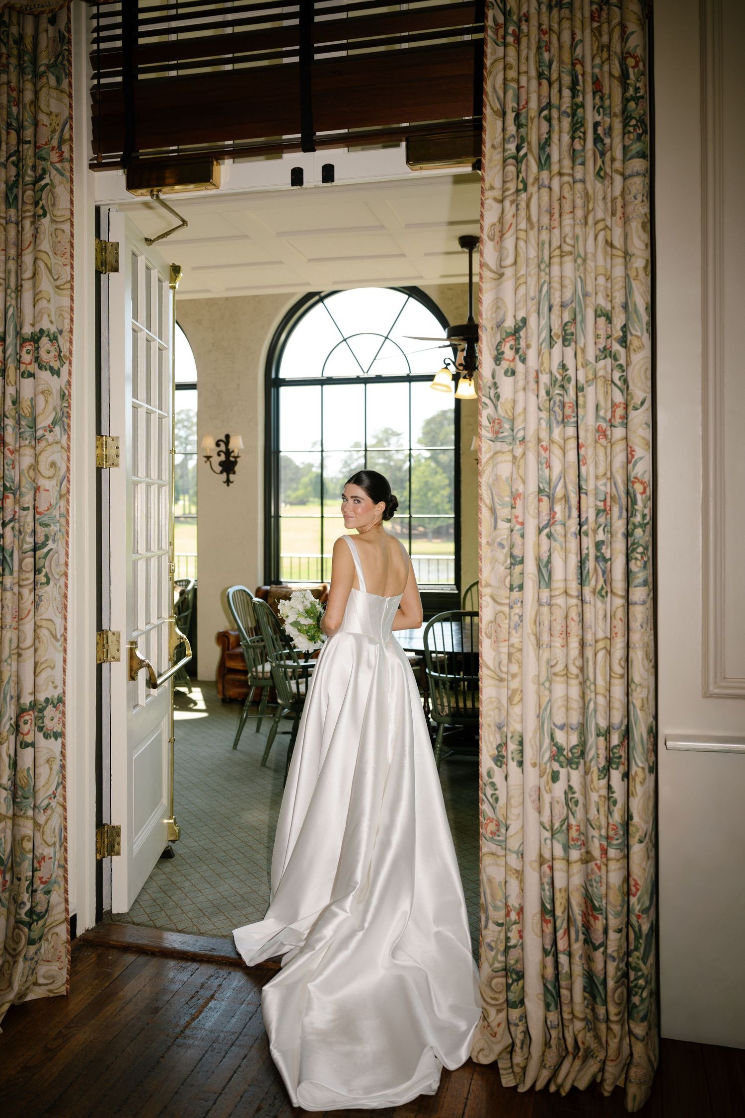 Elegant bride in a white satin gown standing in a doorway with floral curtains, gazing toward an arched window.