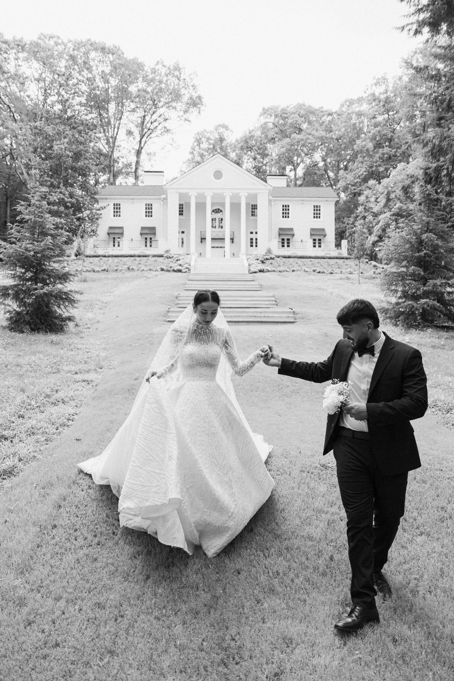 A bride and groom dance playfully across a grassy lawn with a stately white mansion in the background.