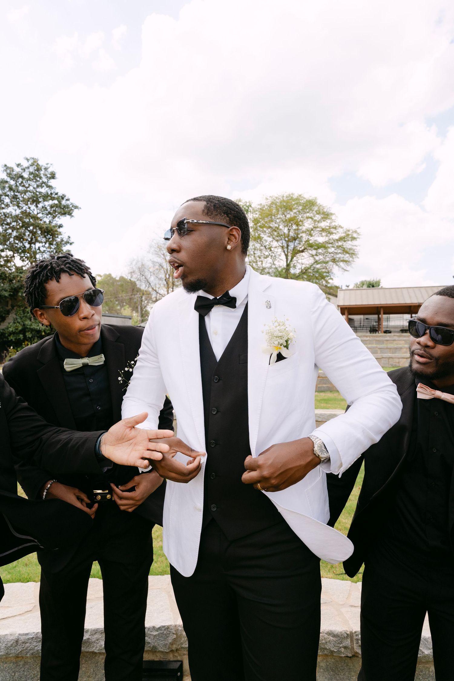Groomsmen in formal attire share a candid moment outdoors, with the groom wearing a striking white jacket.