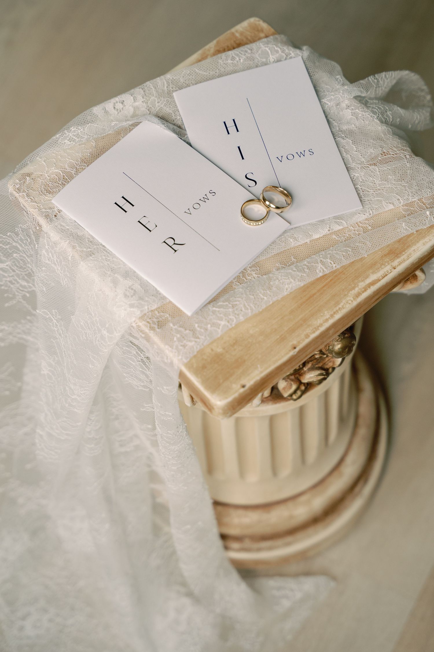 Wedding rings and vows cards rest on an elegant white lace fabric and decorative column pedestal.